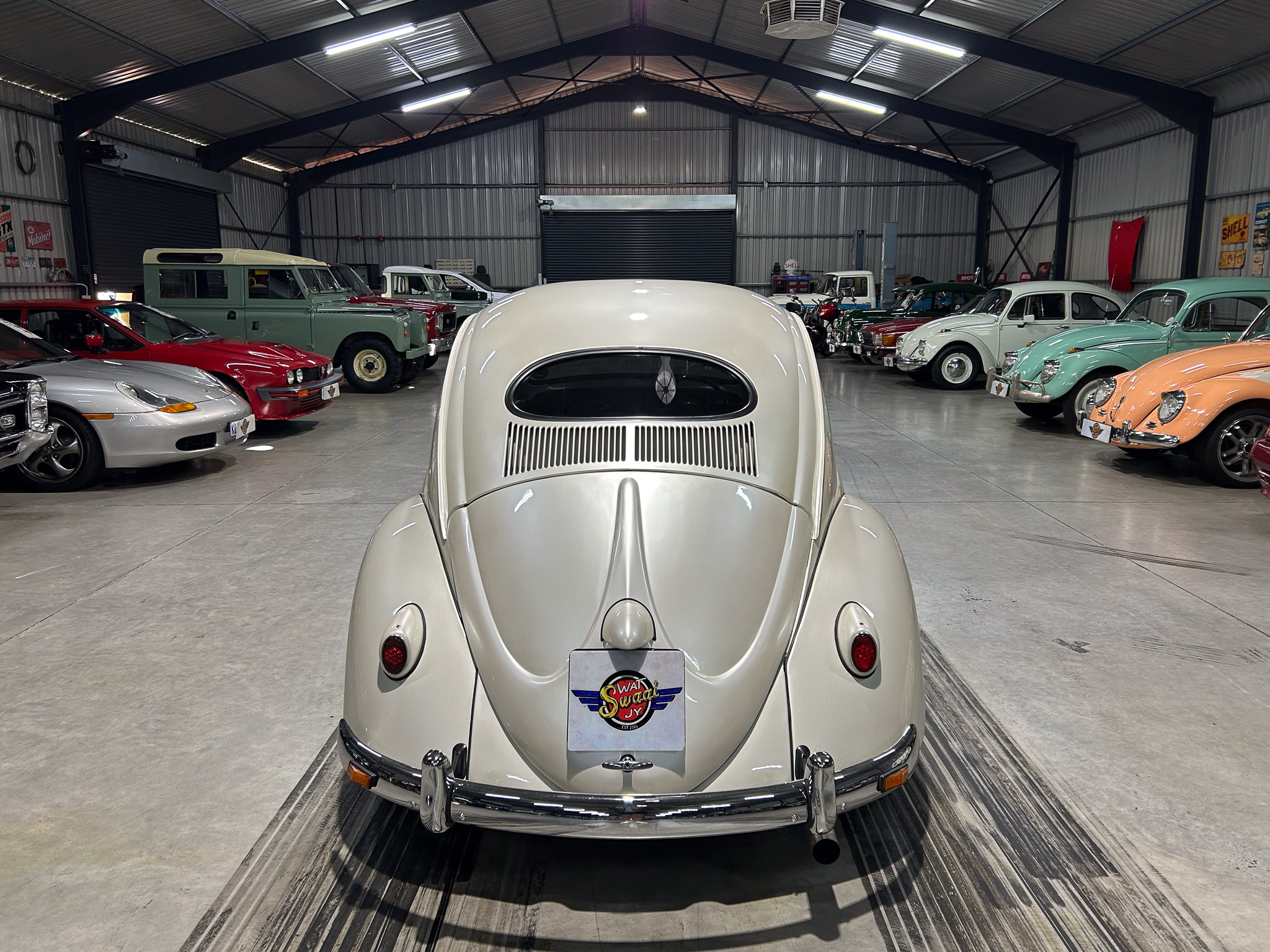 Front and center view of a vintage white car in a spacious warehouse showroom filled with classic cars on both sides.