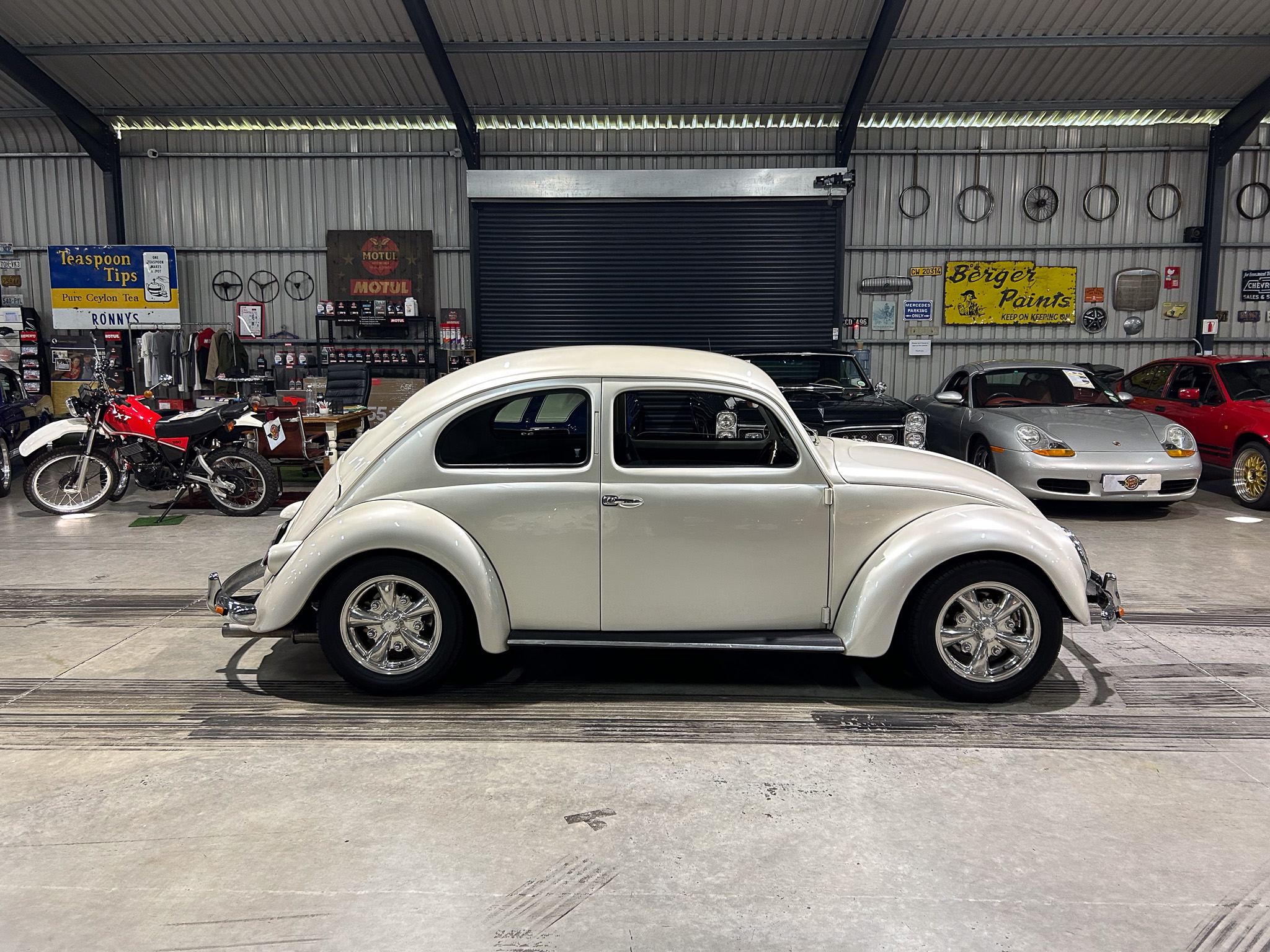 White vintage car in a showroom foreground, with other classic cars, a red motorcycle on the left, and retro metal signage on the walls