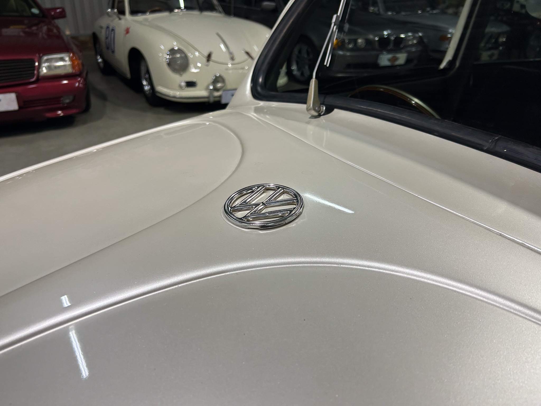 Close-up of a pale vintage car hood with a chrome emblem and antenna, indoors at a car showroom with other classic cars in the background.