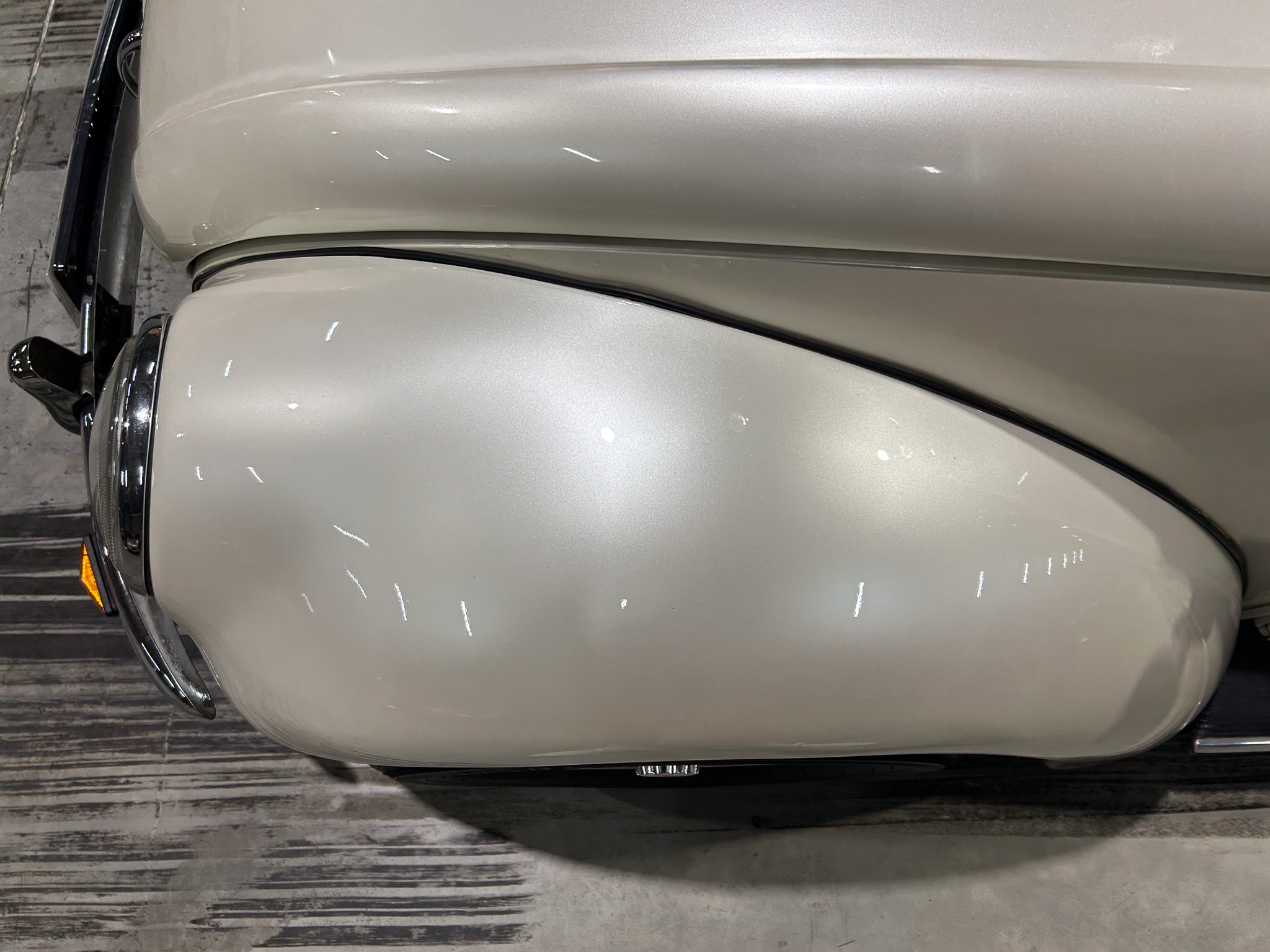 Close-up of a silver vintage car’s front fender and hood, with chrome bumper and an orange turn signal, indoors on a wooden floor.