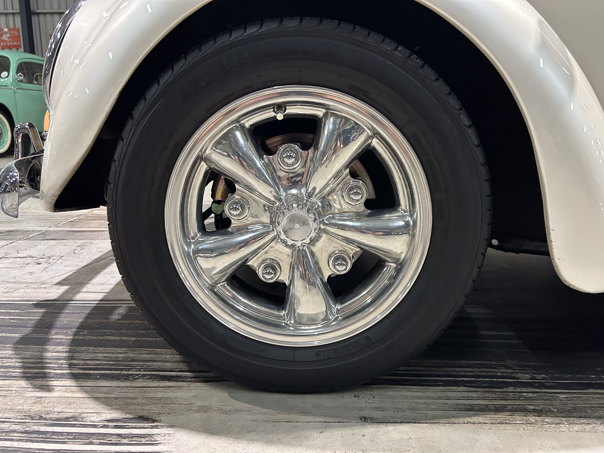 Close-up of a chrome wheel and tire on a white vintage car, with a green car in the background.