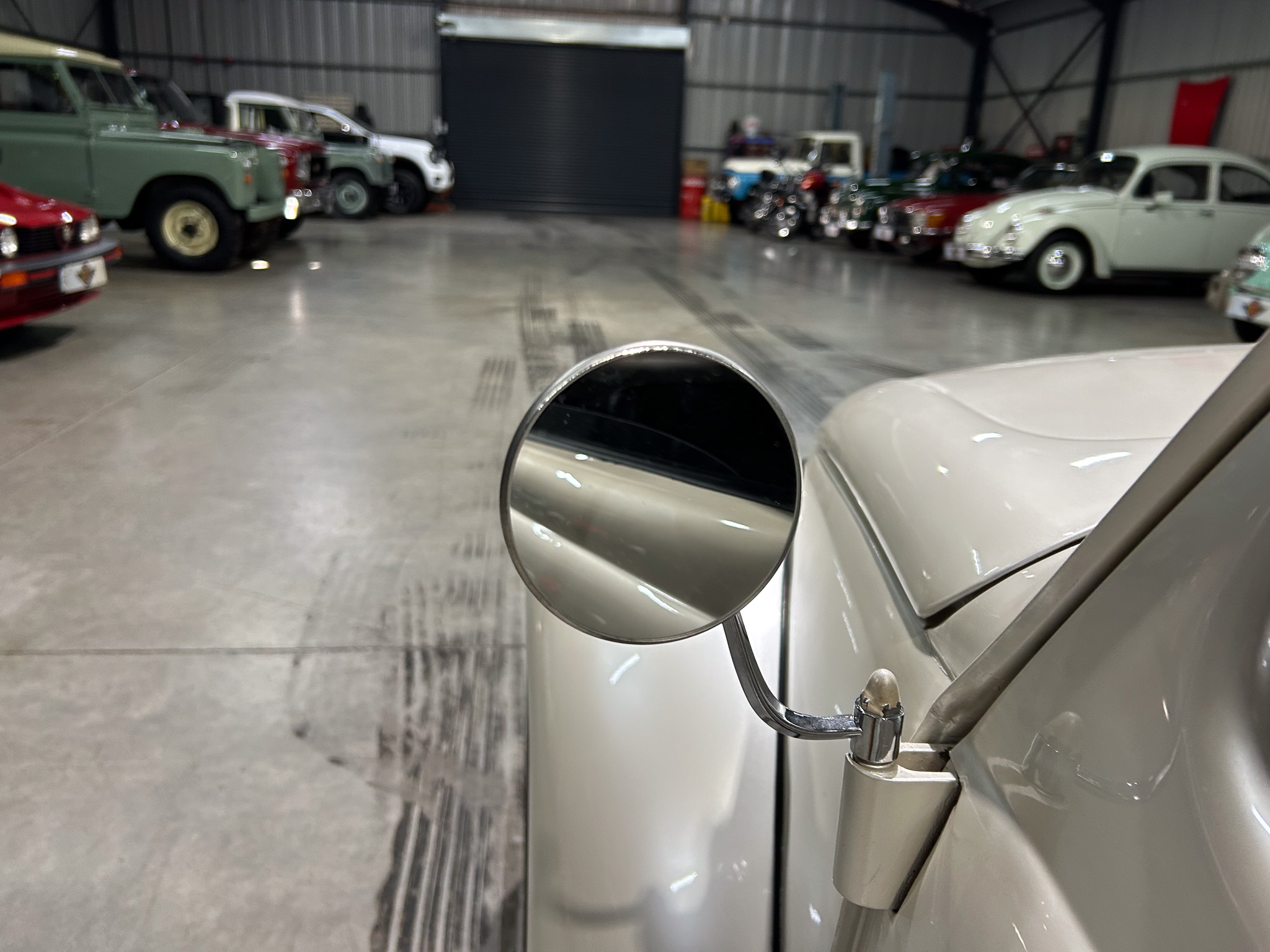 Close-up of a car's round side mirror with a row of vintage cars in a dim showroom background, metal floor visible behind.