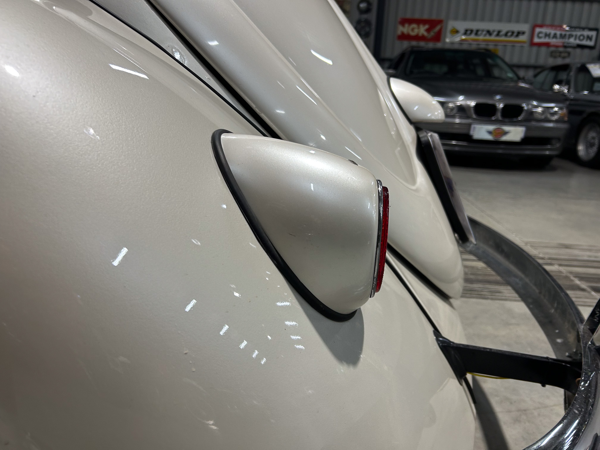Close-up of a cream-colored vintage car's rear fender with chrome trim in a showroom. In the background, another classic car and dealership banners are visible.