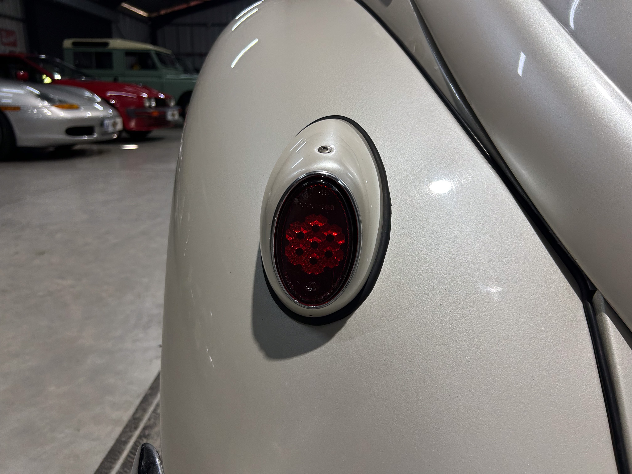Close-up of a vintage silver car's round red taillight on a smooth body, with a garage and other cars blurred in the background.