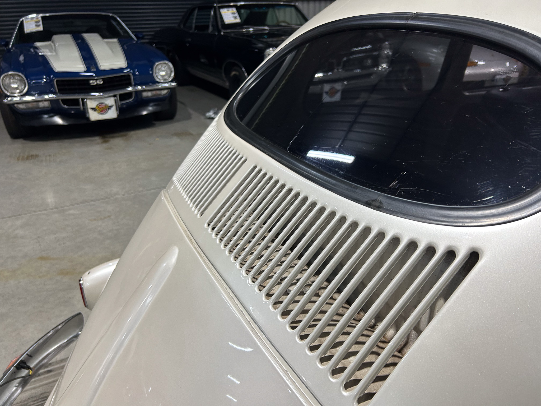 Close-up of a cream-colored vintage Porsche 911 rear engine deck with vent slats; a blue muscle car is visible in the background in a showroom.