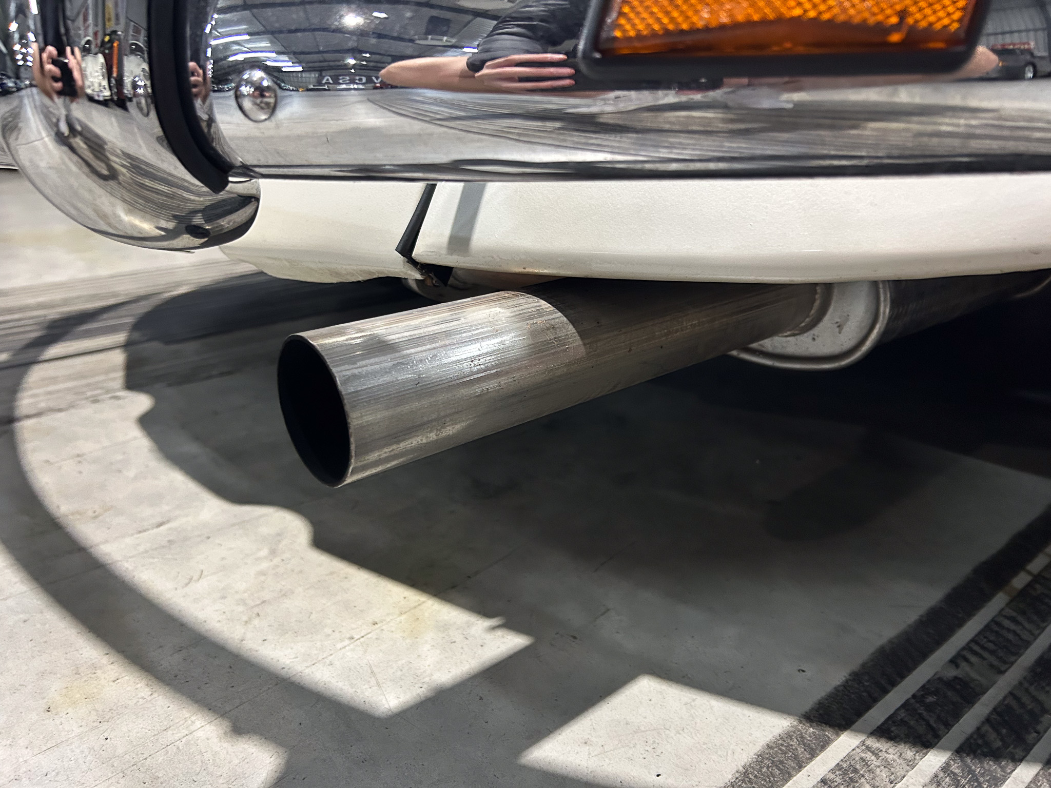 Close-up of a car's chrome bumper with two exhaust tips visible beneath and an orange rear light nearby.