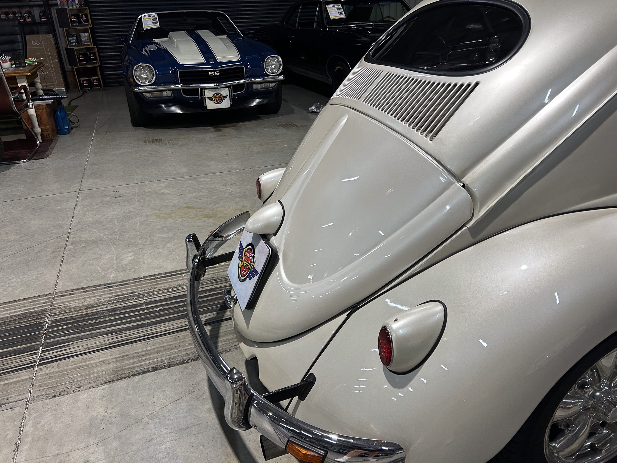 Indoor garage scene with two classic cars: a light gray Porsche viewed from the rear in the foreground and a blue Chevrolet Camaro SS with white racing stripes parked in the background behind it.