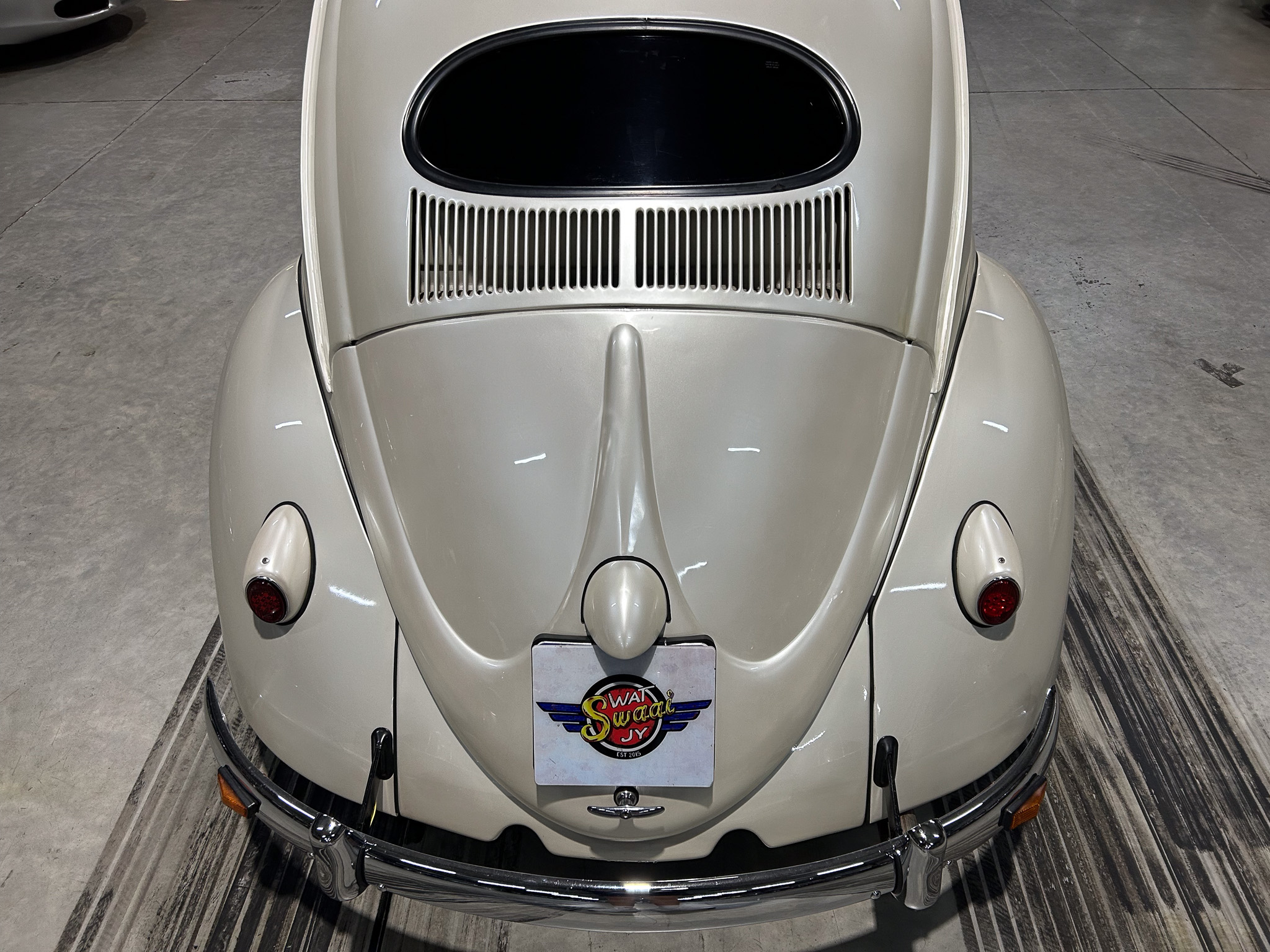 Rear view of a light-gray vintage car showing the rounded fenders, chrome bumper, and vertical grille slats beneath a small rear window; license plate area displays a colorful 'Swat Swag' badge/graphic.