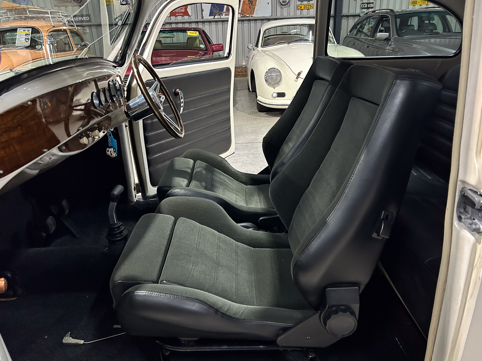 Open car door reveals vintage interior: wooden dashboard, round gauges, and dark gray seats with a steering wheel in view, plus a white classic car parked outside.