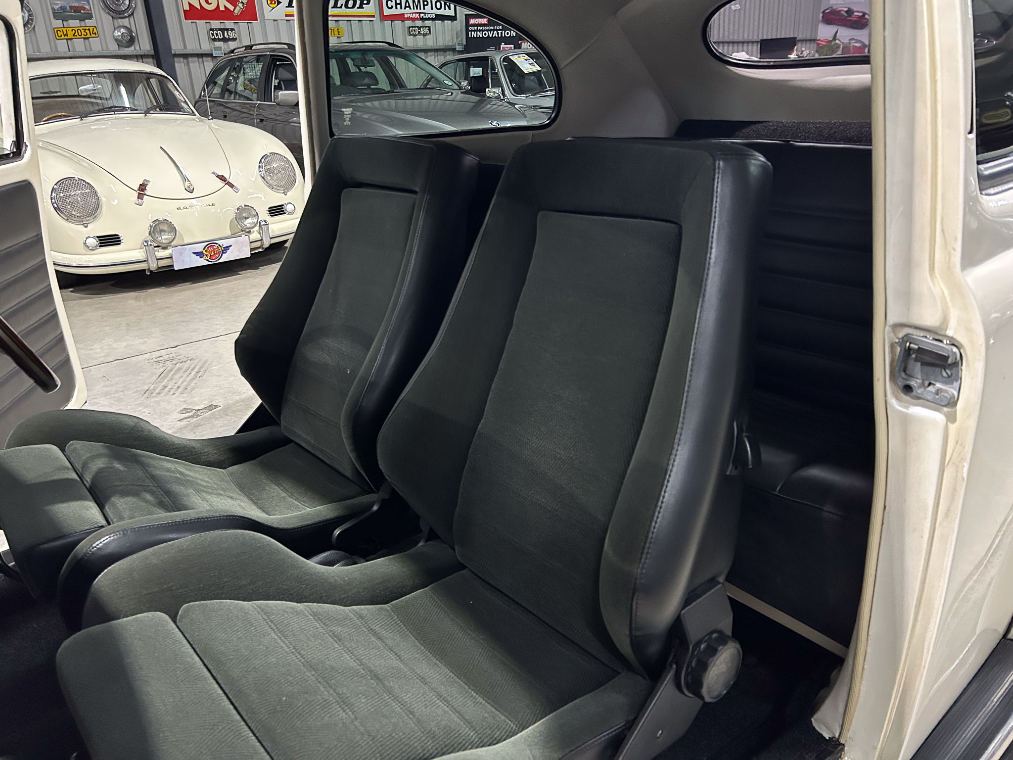 Interior of a vintage car with black bucket seats; open door reveals a white classic car in a showroom behind.