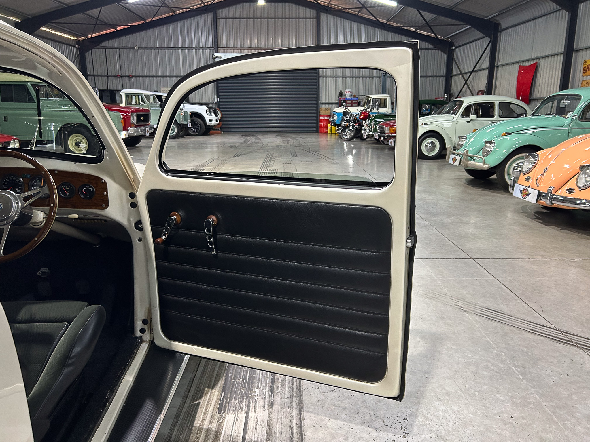 View from the driver's seat of a vintage car, open door revealing a classic showroom filled with pastel-colored cars and motorcycles in the background