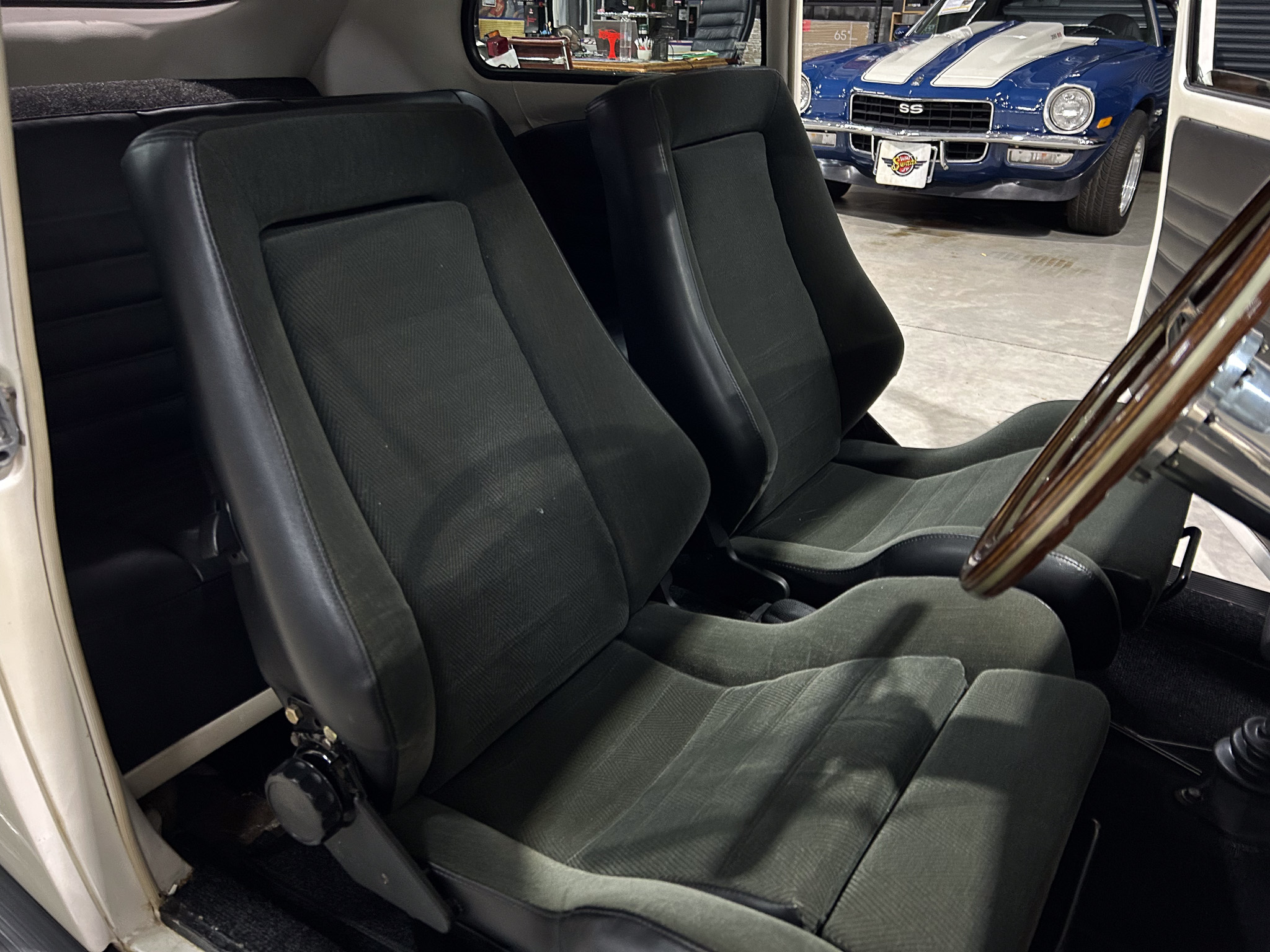 Interior of a vintage car with black cloth seats; open door reveals a blue classic car in a showroom.