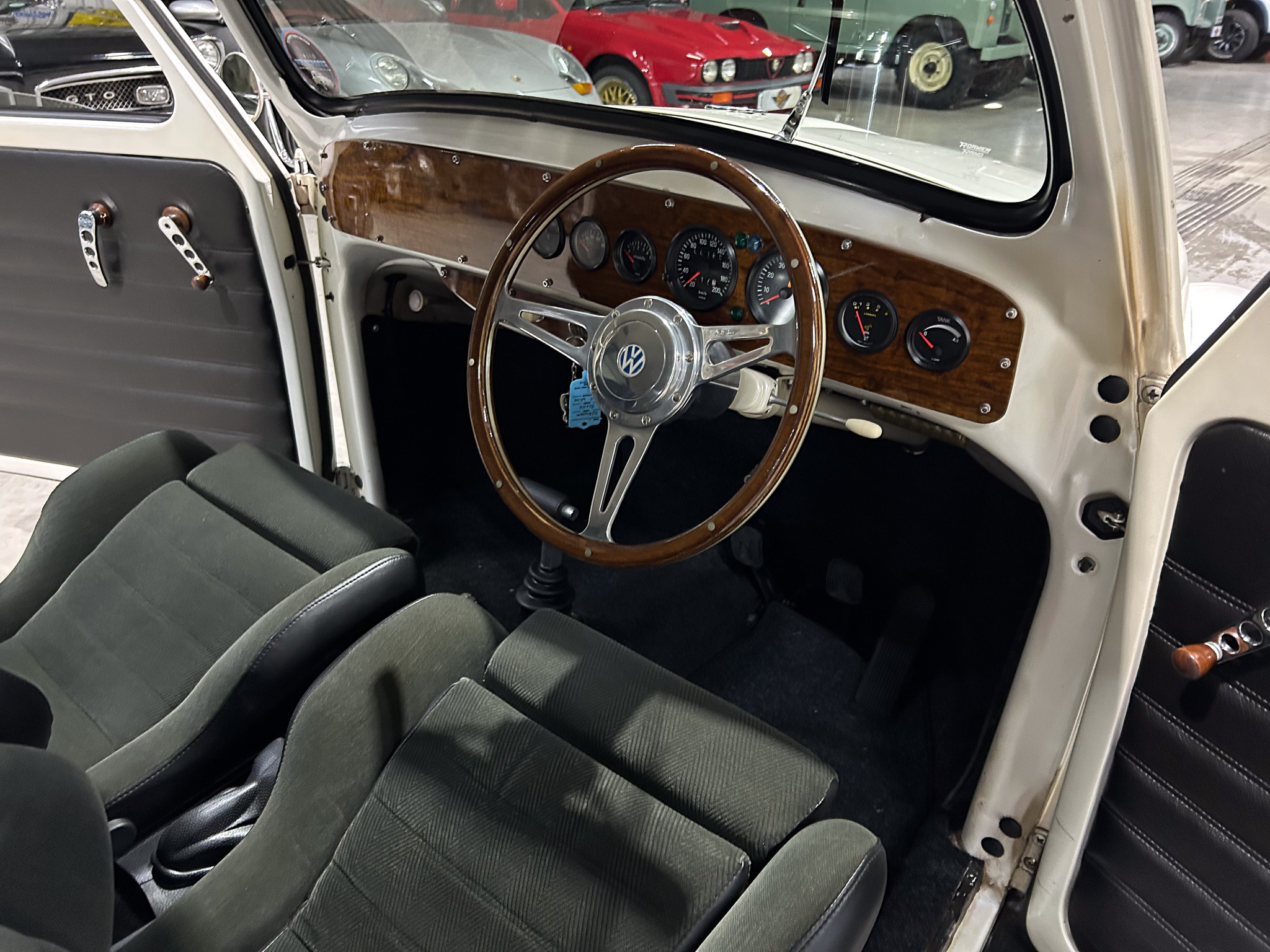 Interior of a vintage car: wood-grain dashboard, multiple analog gauges, and a polished wooden steering wheel with chrome spokes; door open on the left.