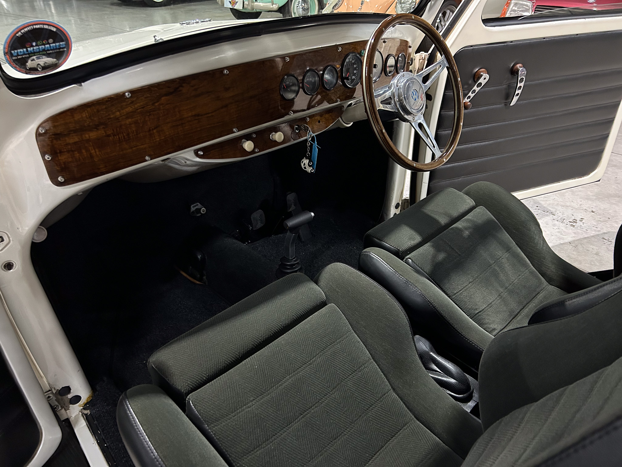 Interior of a vintage car showing a wood dash with multiple gauges and a wooden steering wheel. Grey door panels and fabric seats are visible.