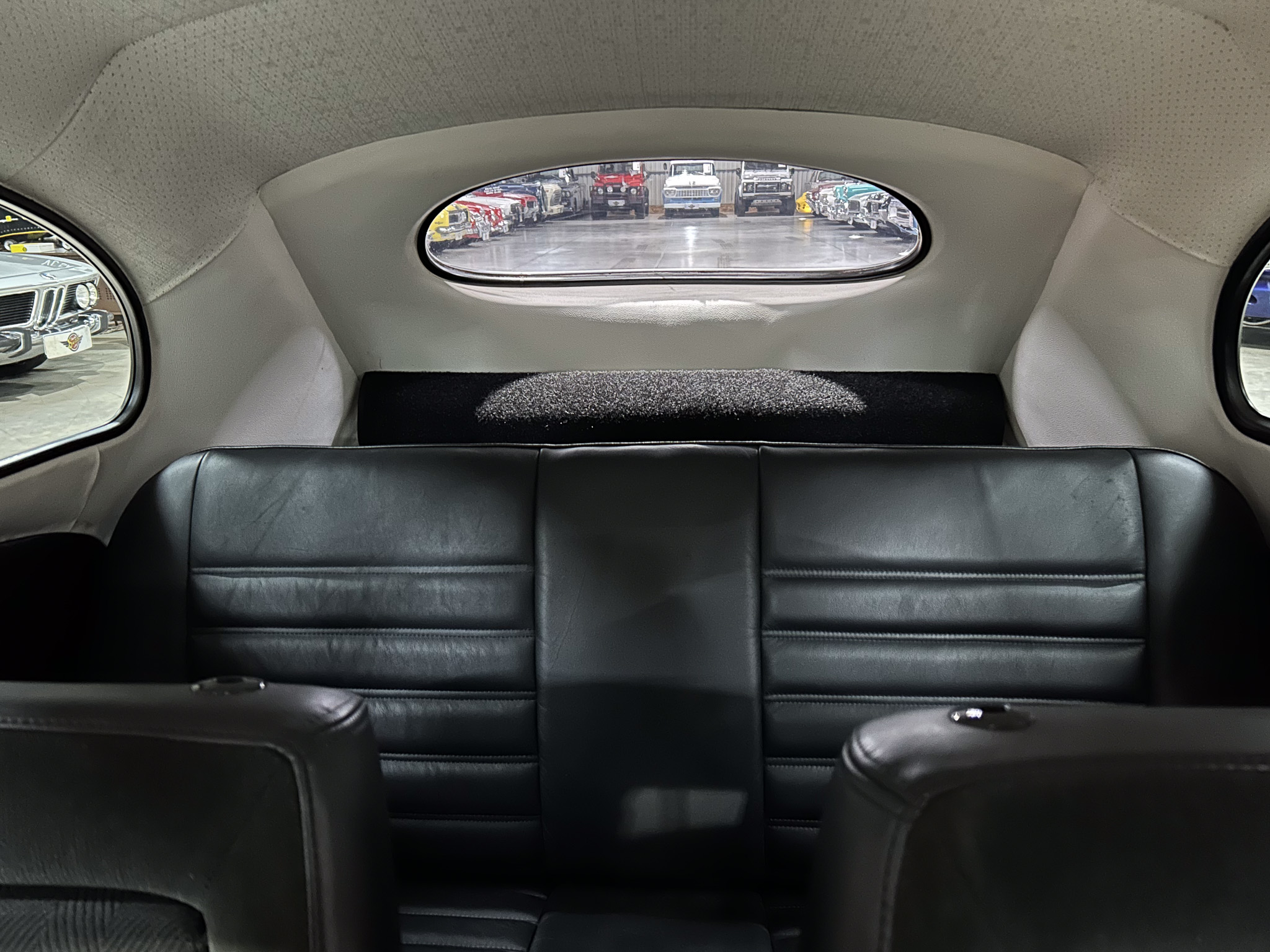 View from the back seat of a car looking through an oval rear window at a row of vintage cars parked in a showroom/garage.