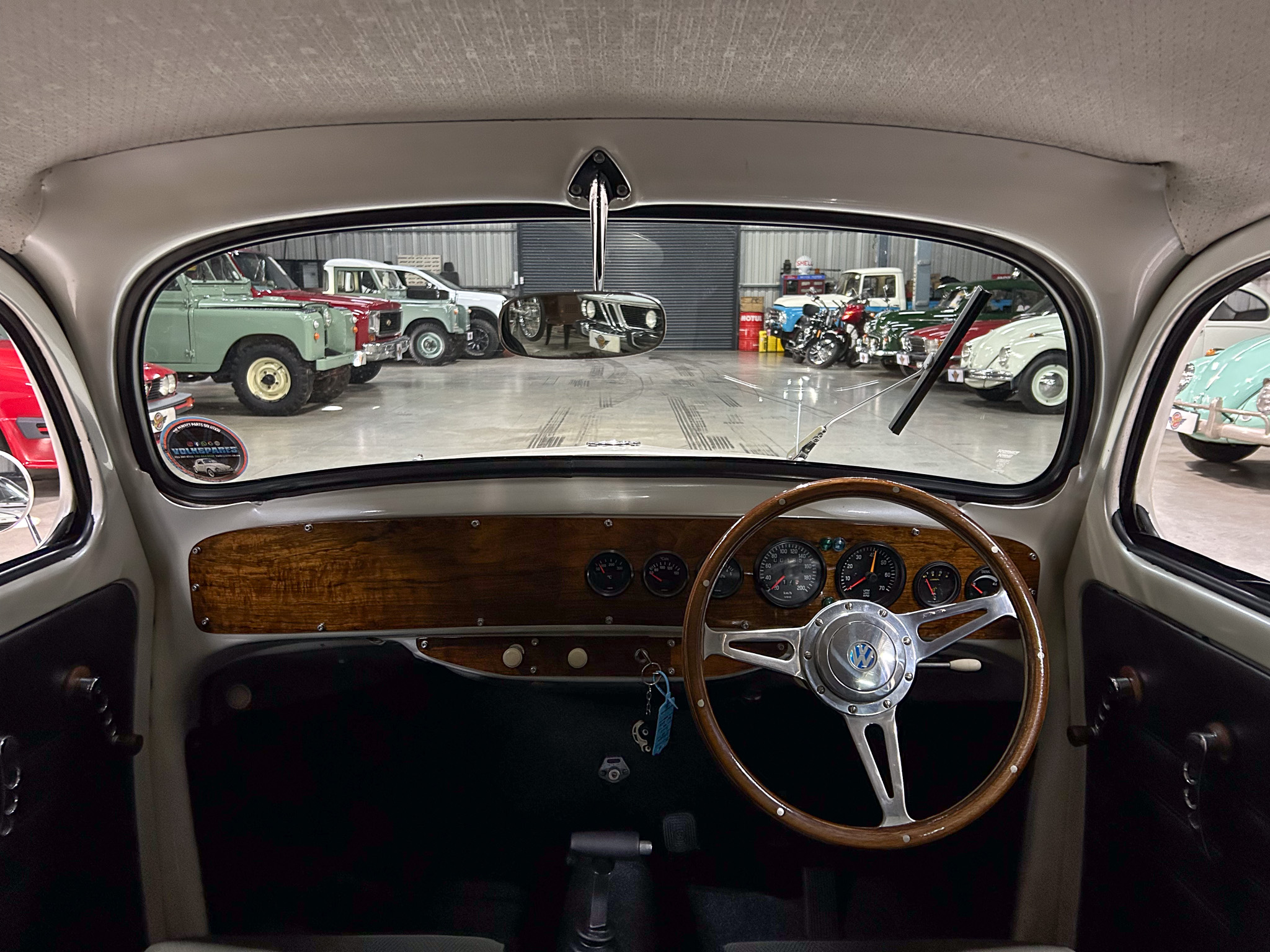 Interior of a vintage car with a wooden dashboard and classic steering wheel, looking out at an auto museum filled with antique cars