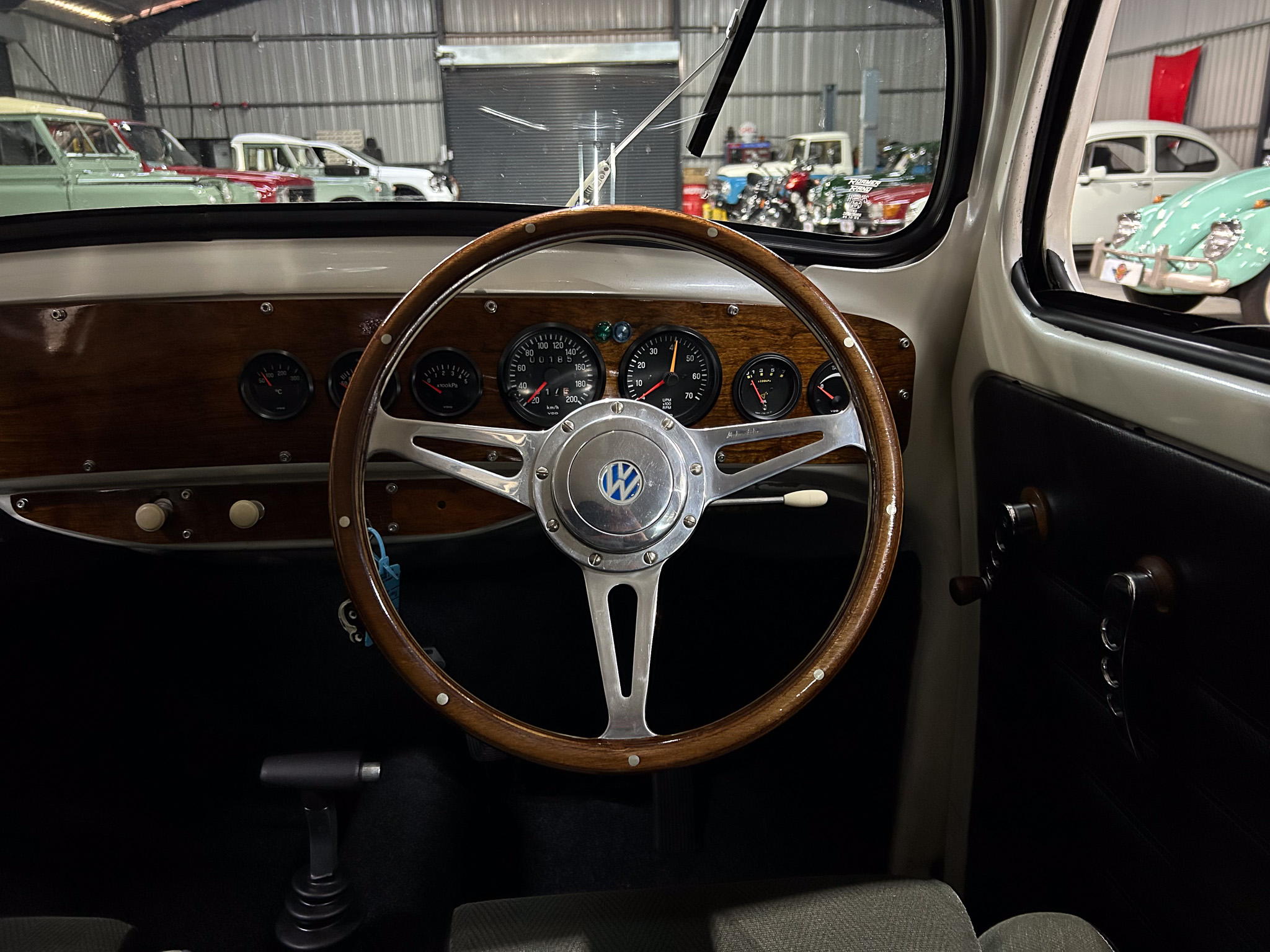 Vintage car interior: wood dashboard, metal steering wheel with VW emblem, classic gauges visible through the wheel. Outside the workshop show vintage cars.
