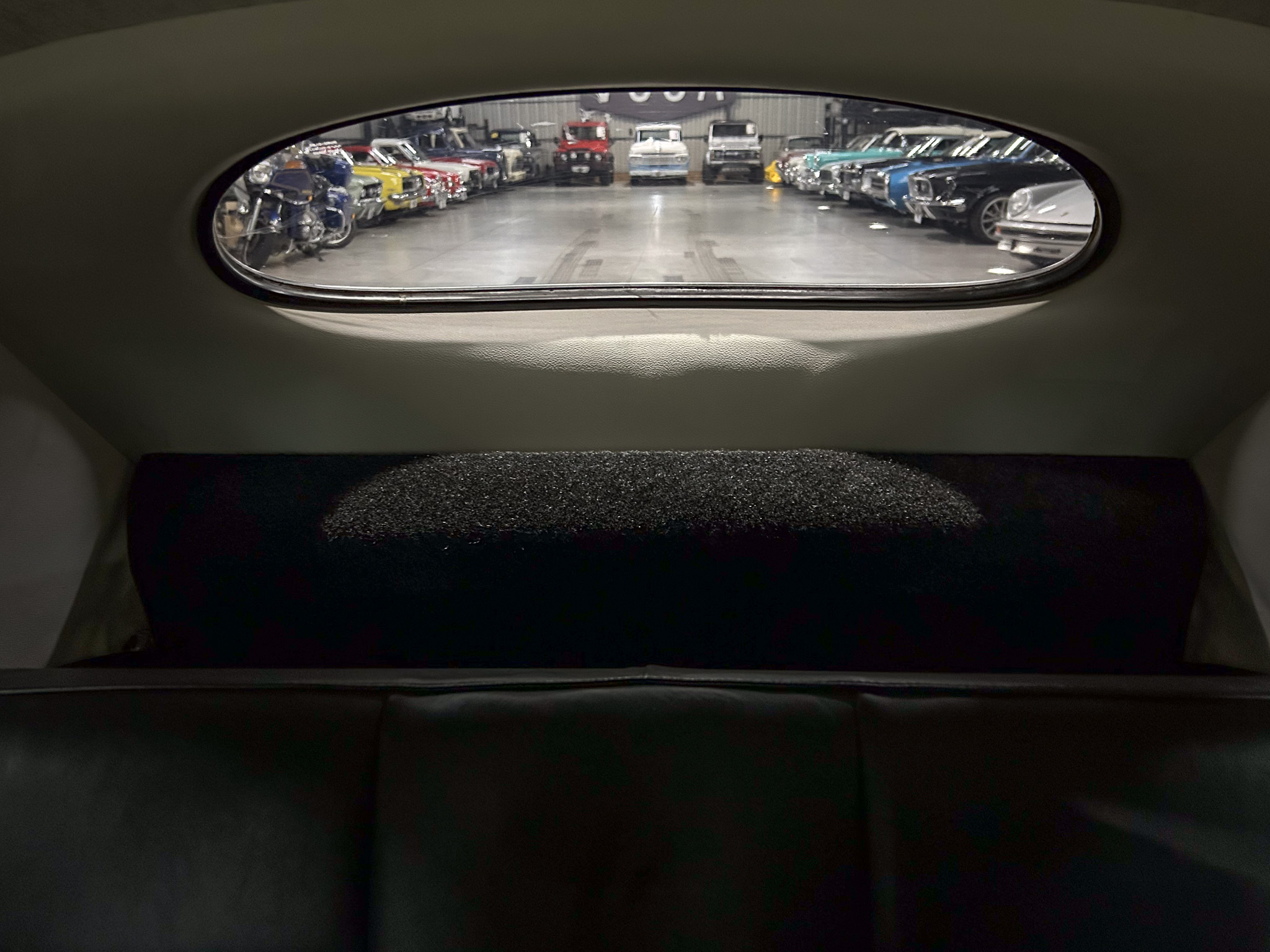 View through a car's rear window into a showroom filled with vintage cars lined up along both sides of an open space at the center of the aisle.