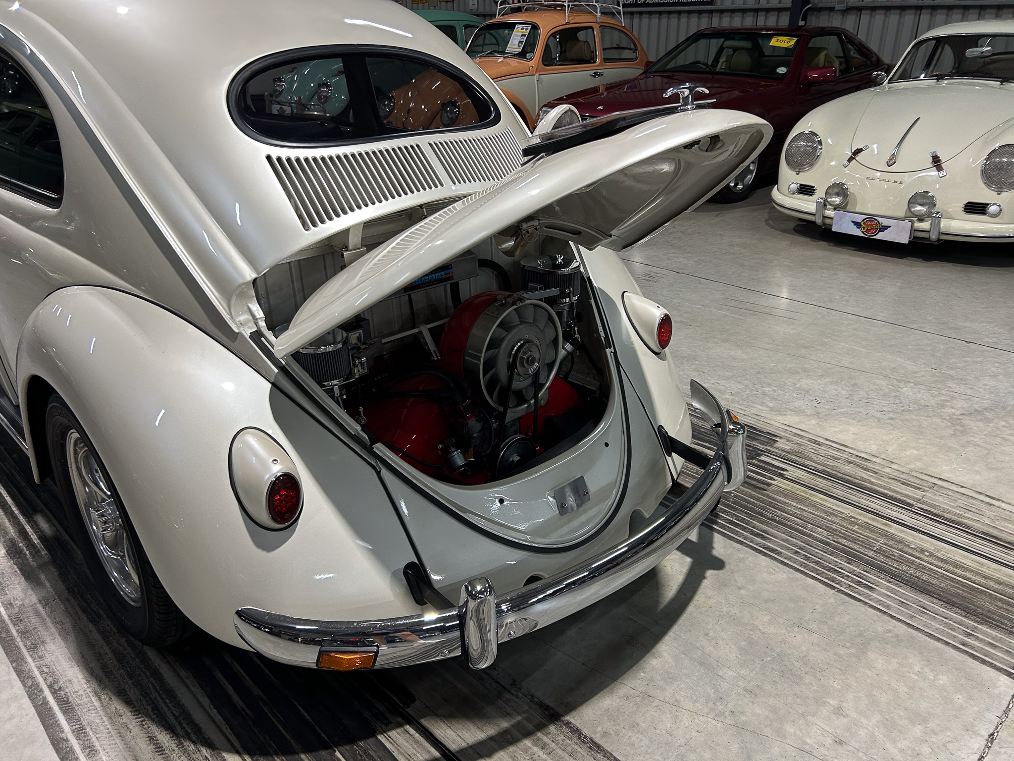 White vintage Porsche 356 with rear hood raised, exposing the red engine components in a classic car showroom.