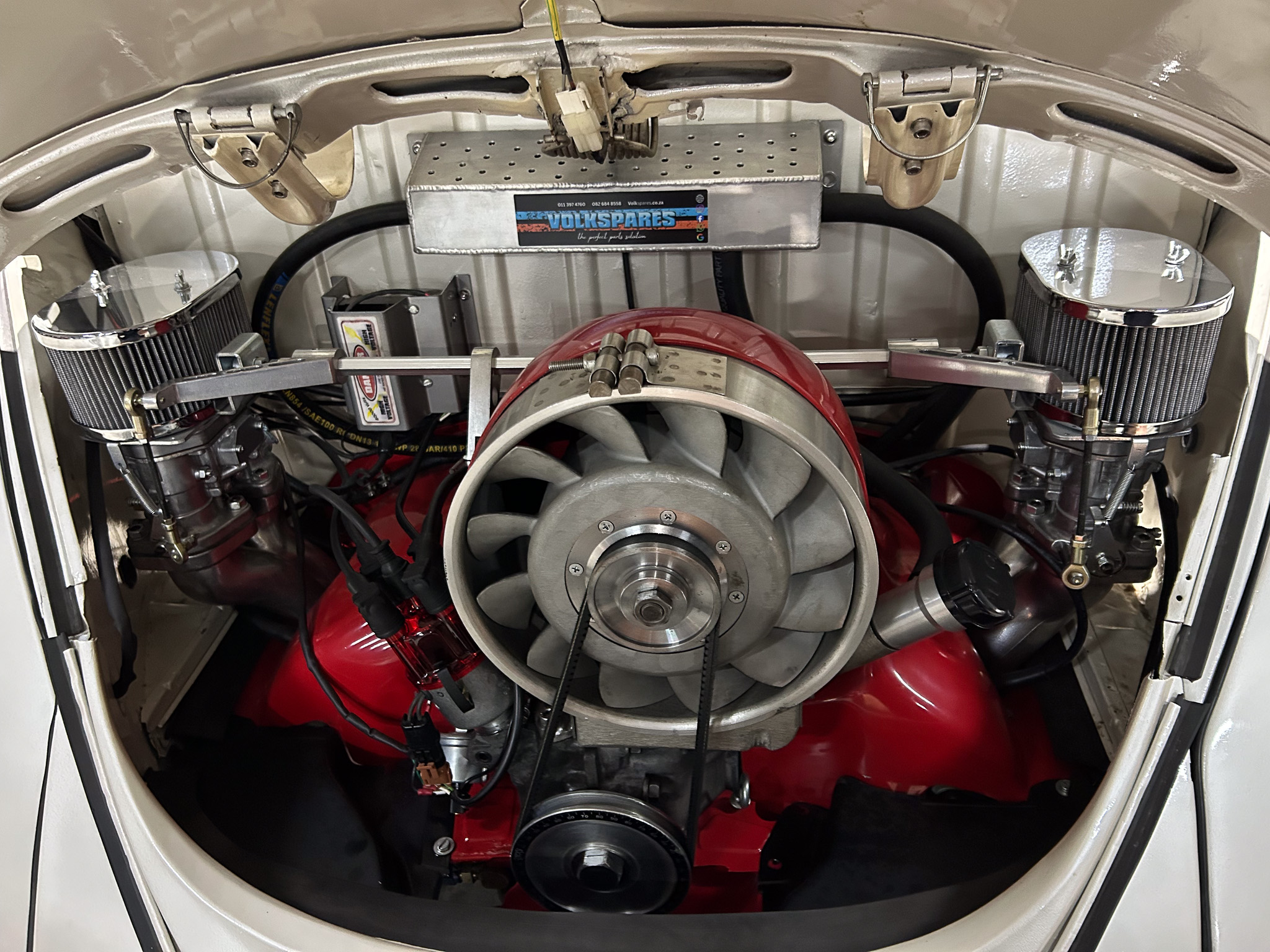 Open engine bay of a race car showing dual chrome air filters, belts, and wiring with a red fire-suppressant bottle nearby