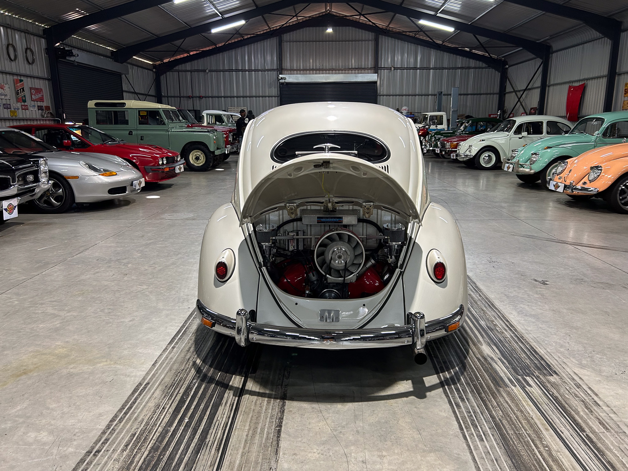 Beige vintage car with open hood in a large showroom, surrounded by pastel classic cars in a museum-like garage.