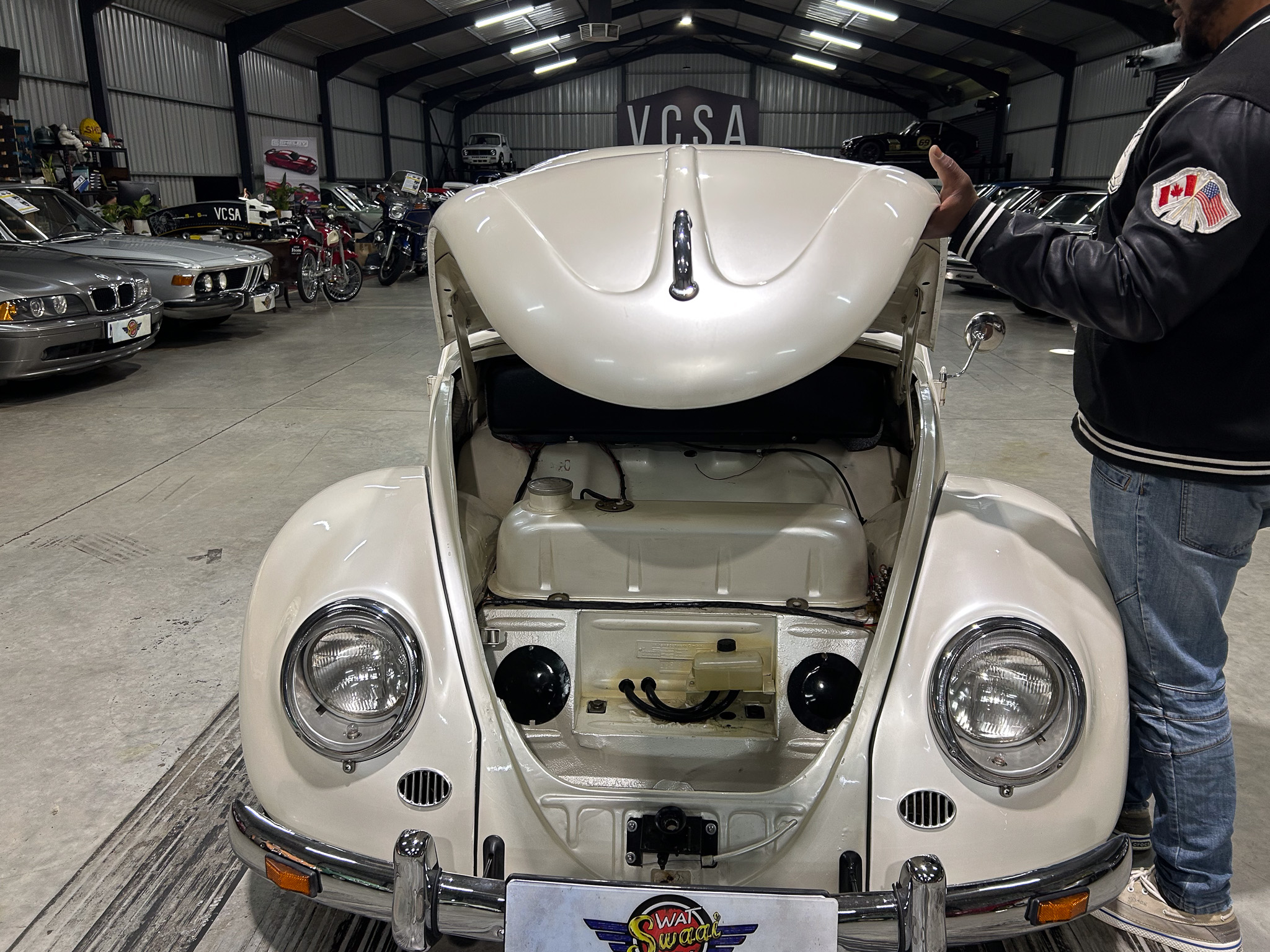 Front view of a white vintage car with its hood open in a spacious garage, a man in a leather jacket stands to the right.