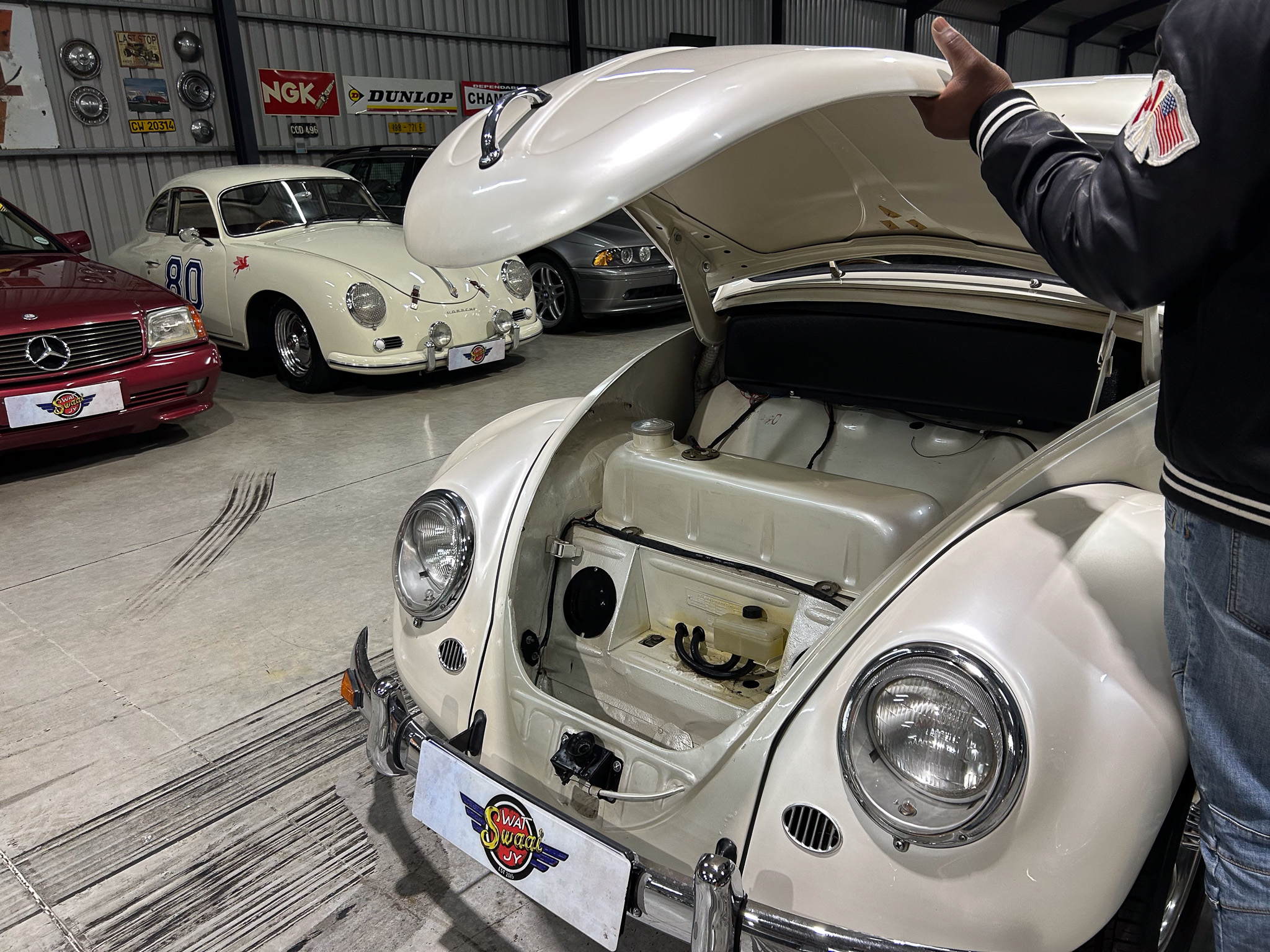 White vintage car with its hood open in a showroom, other classic cars visible in the background, and a person in a dark jacket with an American flag patch standing at the open trunk.