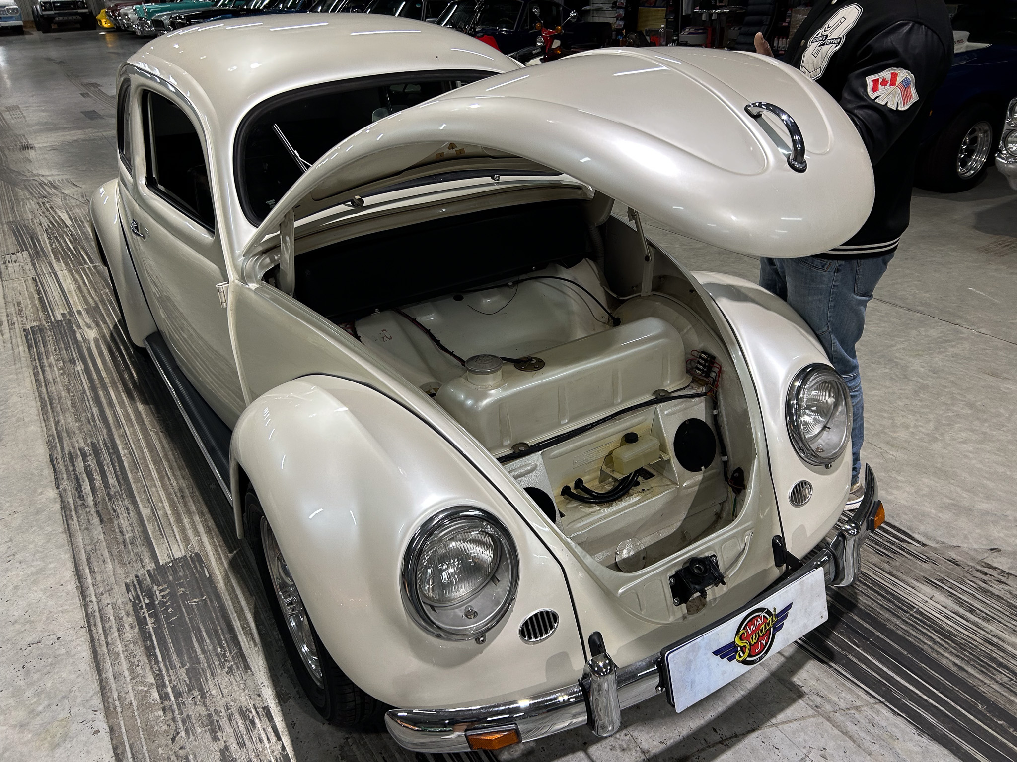 Cream-colored vintage car with its trunk open in a showroom, a person in a patched jacket standing nearby.