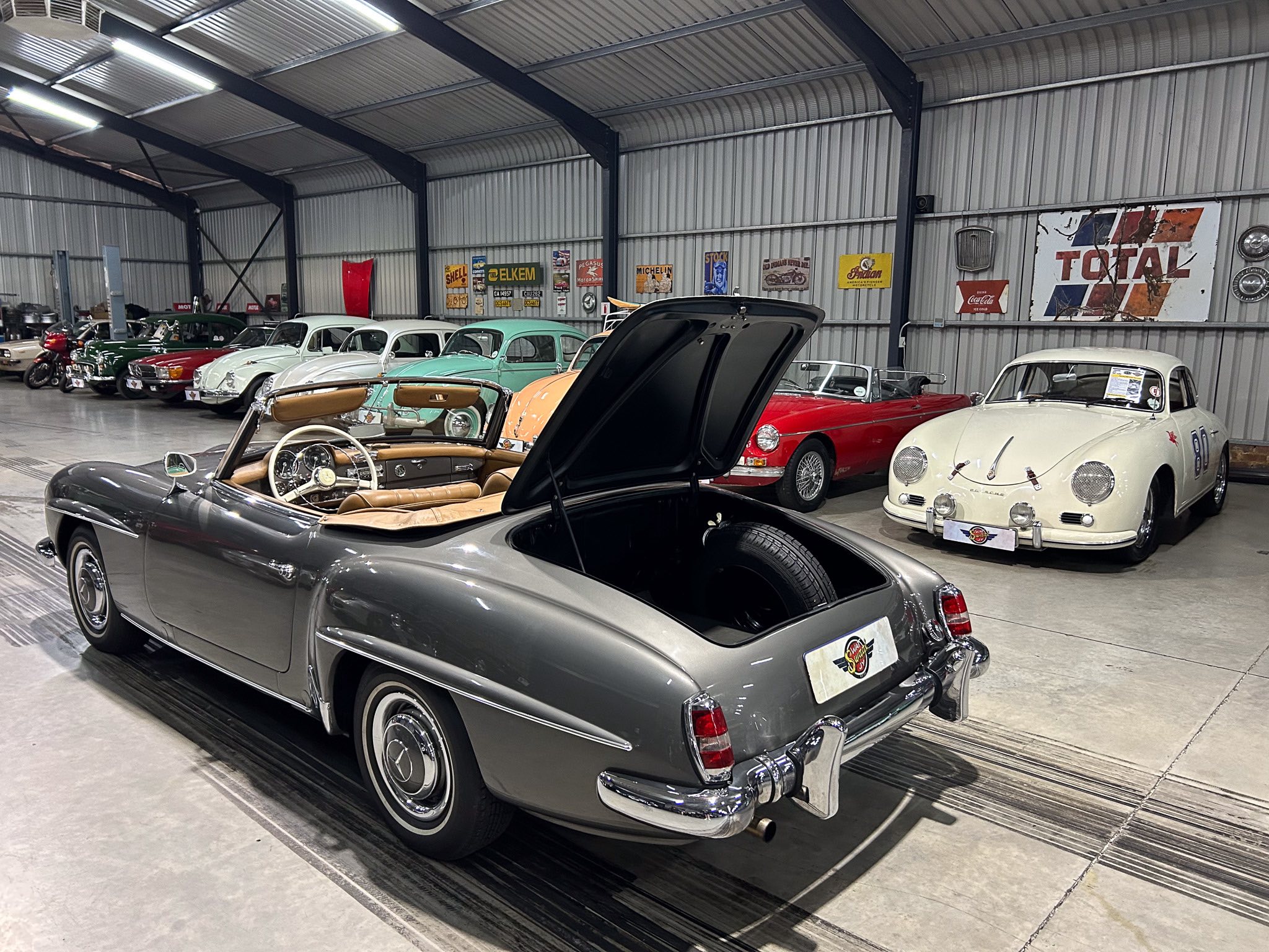 Indoor showroom filled with vintage cars, several classic convertibles and sedans on display against corrugated metal walls with retro signage