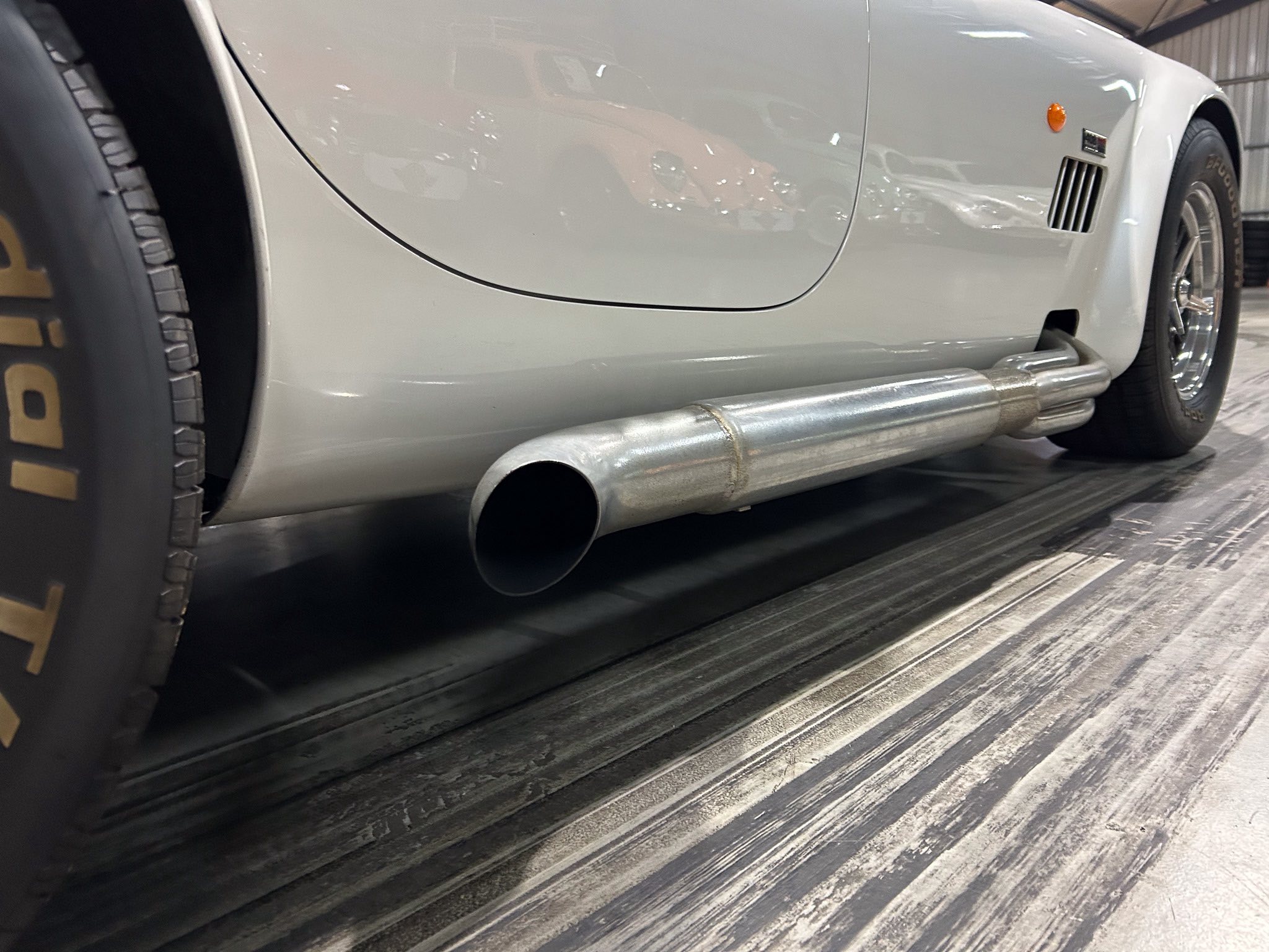 Side view of a silver classic car showing an exposed exhaust pipe and rear wheel in a showroom.