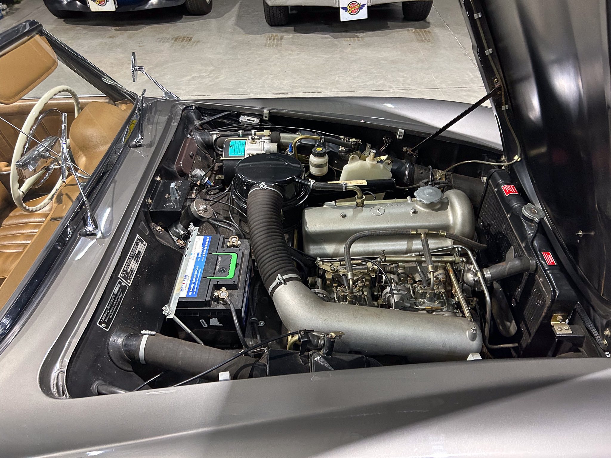 Classic car engine bay with exposed components; tan leather seats and steering wheel visible inside the open cabin, hood raised on a vintage Mercedes.