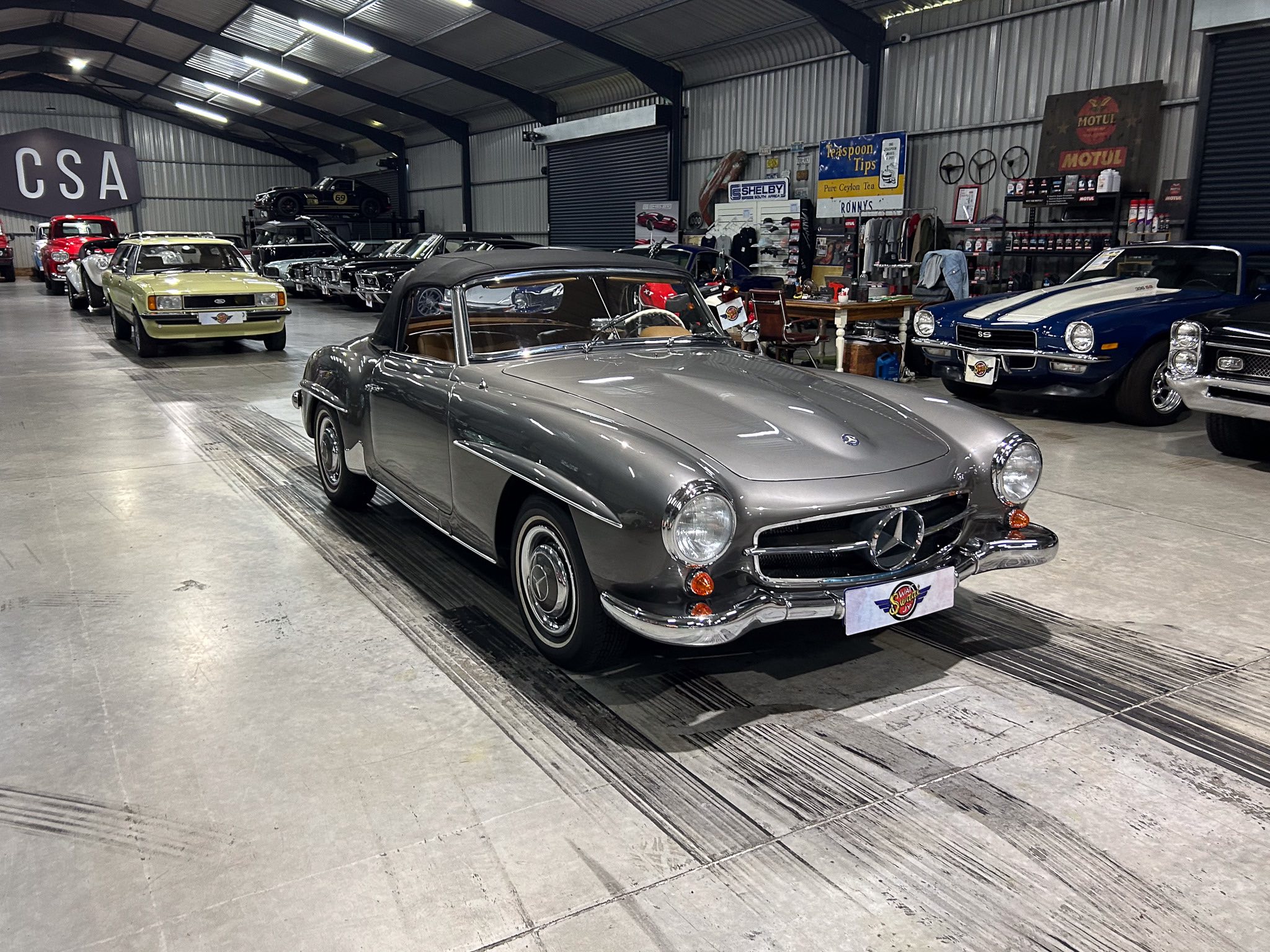 Indoor showroom of vintage cars: a silver convertible in the foreground with other classic cars lined up in a warehouse setting, CSA sign on left.