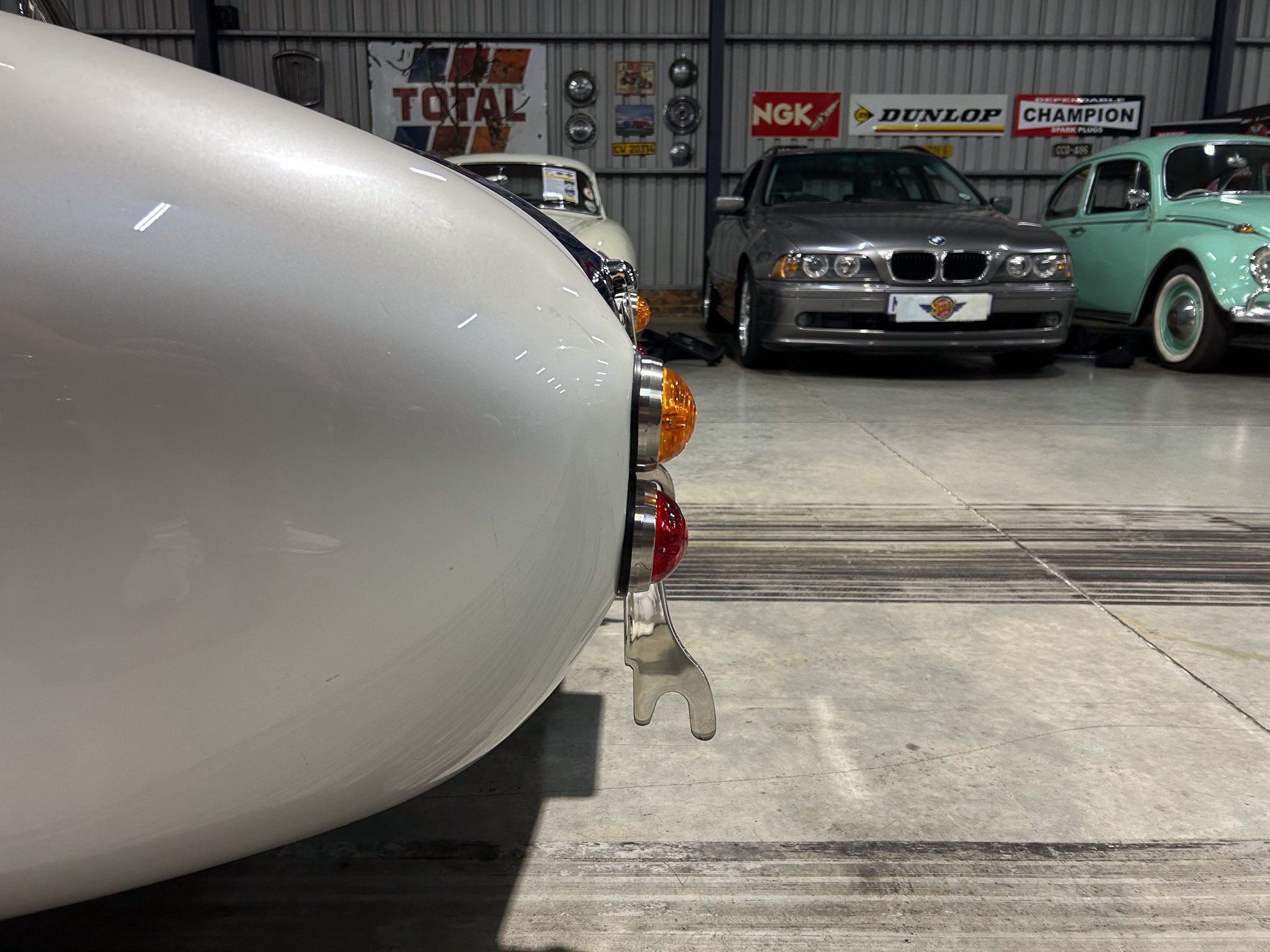 Close-up of the rear left corner of a white vintage car inside a showroom, with other classic cars and retro signage visible in the background.