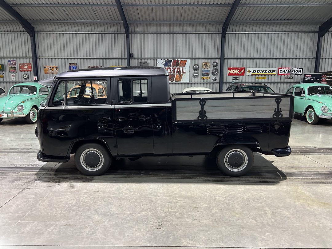 Black vintage pickup with a wooden flatbed, parked indoors among classic cars in a showroom.