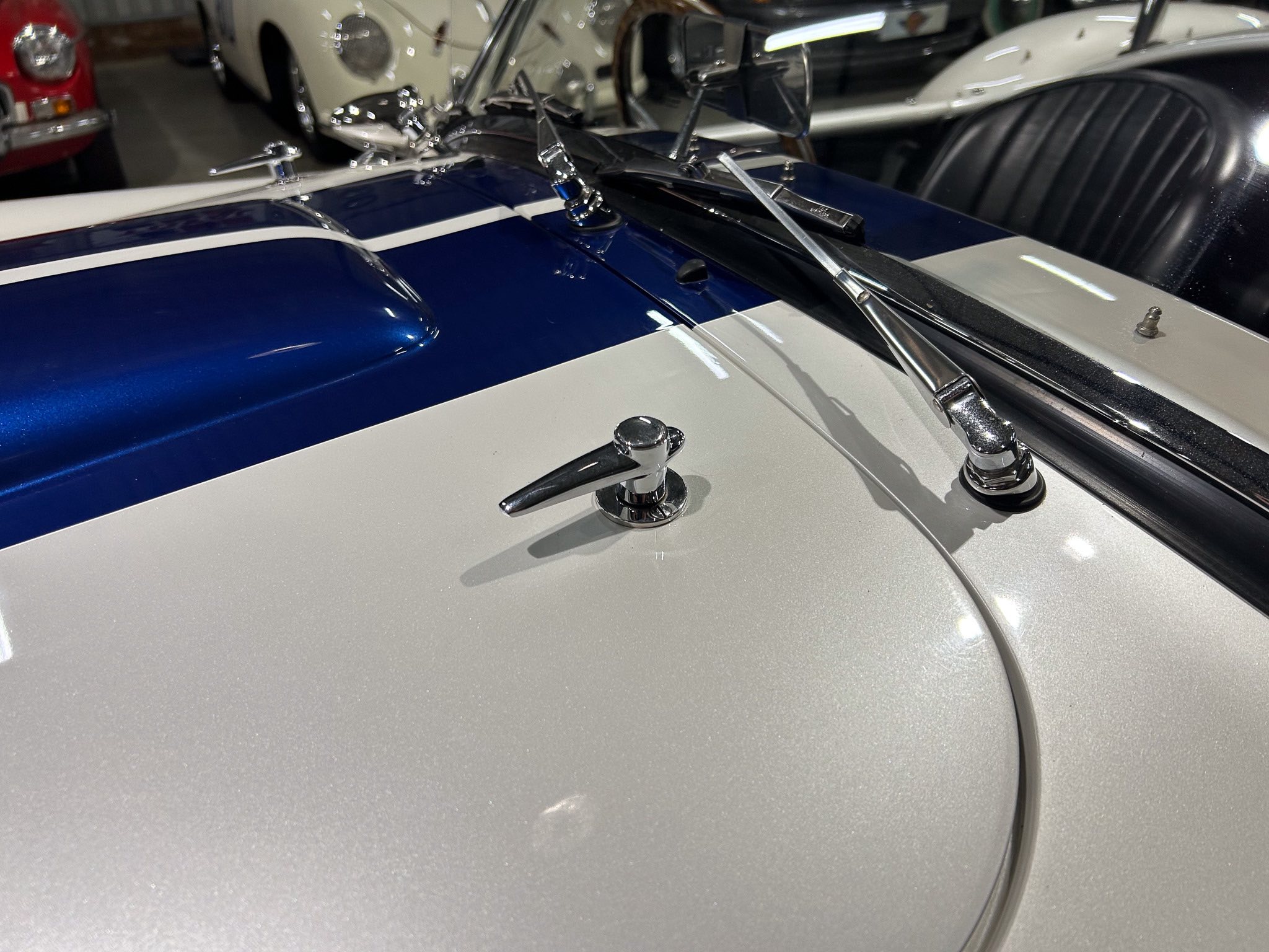 Close-up of a vintage car hood with blue paint, white stripe, and chrome windshield wipers in the foreground.