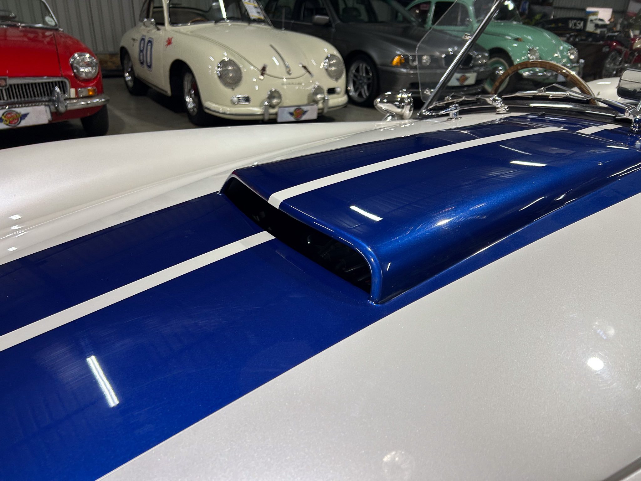 Blue vintage race car with white racing stripe in the foreground; row of classic cars in a showroom behind it.