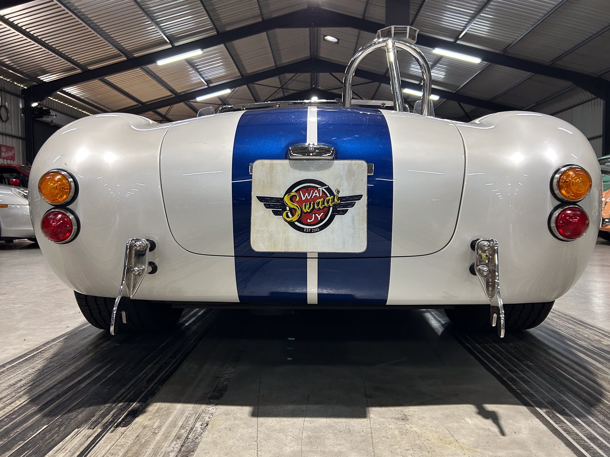 Rear view of a vintage silver sports car with a blue center stripe and chrome bumpers in a garage. Plate area shows a logo badge.