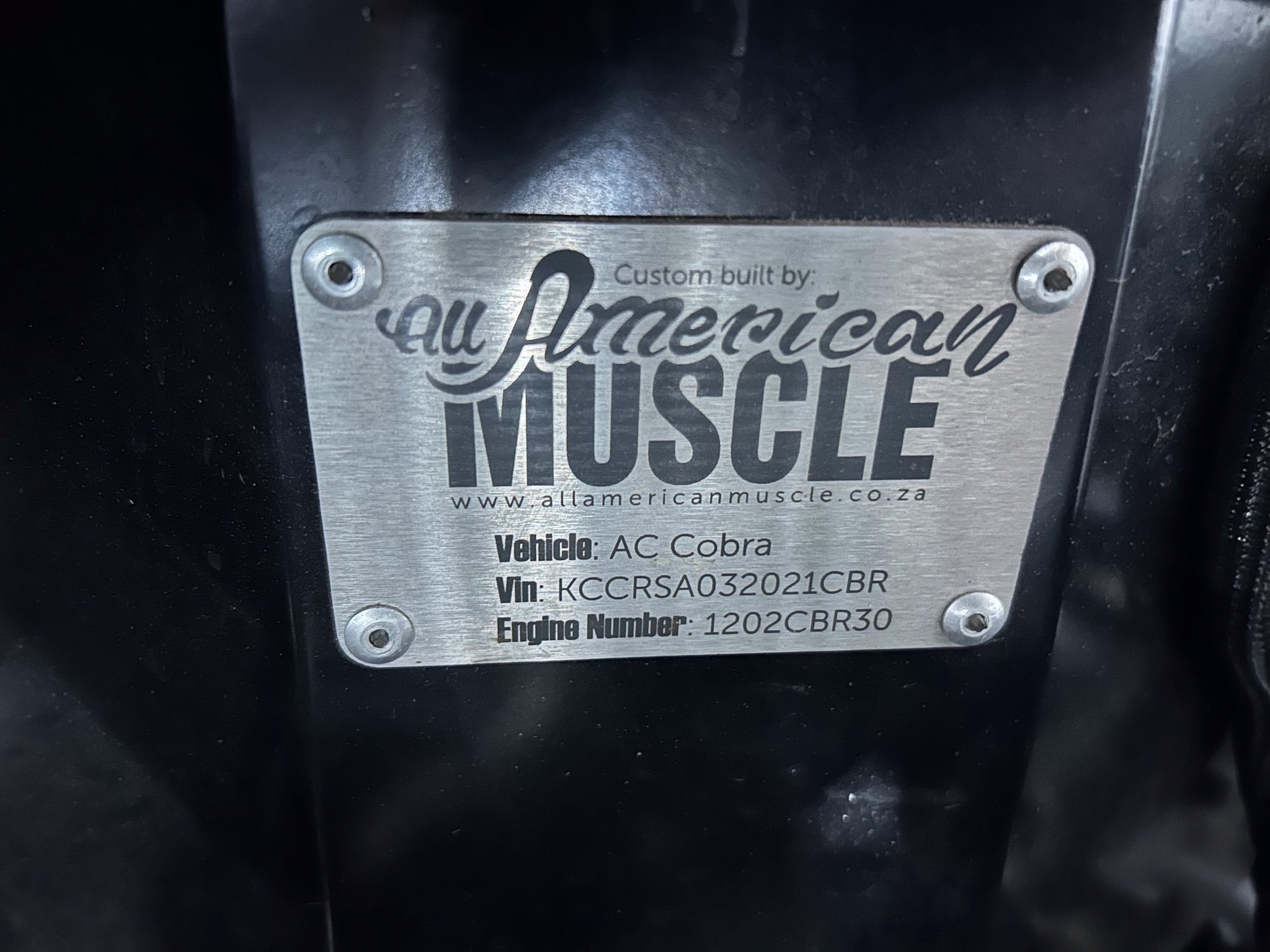 Metal nameplate on a black surface reading 'Custom built by: All American Muscle' with vehicle details like 'Vehicle: AC Cobra' and a web address partially visible, bolted to the panel.