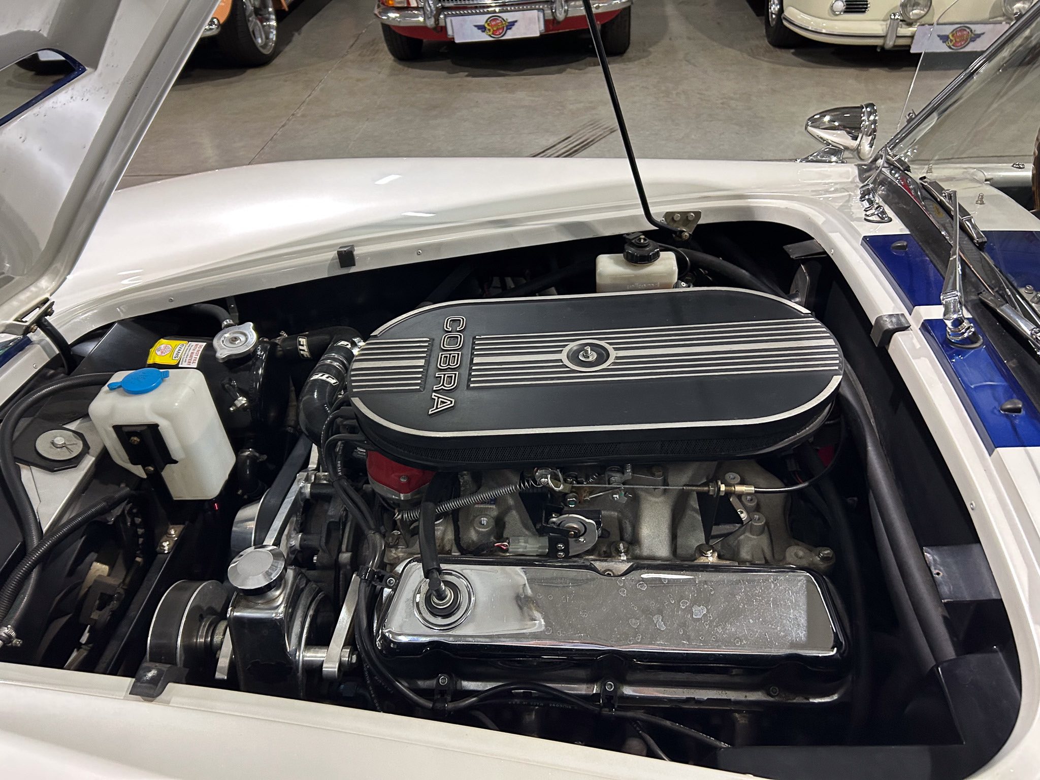 Open hood of a classic Cobra sports car, showing the engine with a 'COBRA' cover and surrounding automotive showroom details.