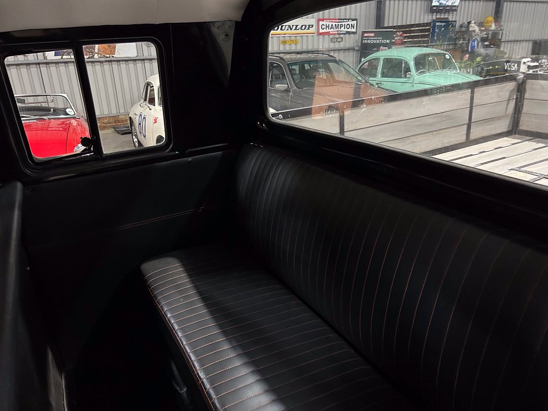 Back seat of a vintage black car with pinstripe upholstery, seen through the rear and side windows at a car showroom or museum yard.