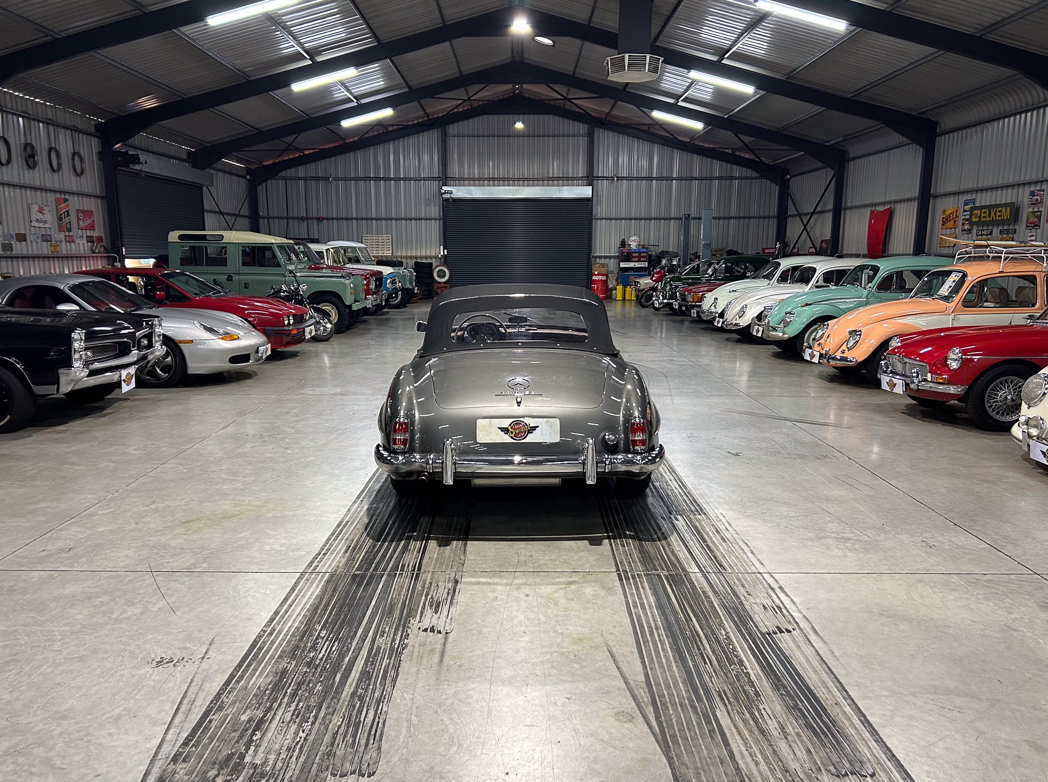 Indoor showroom filled with rows of classic cars lined up along both walls, viewed from the rear of a gray car in the foreground.