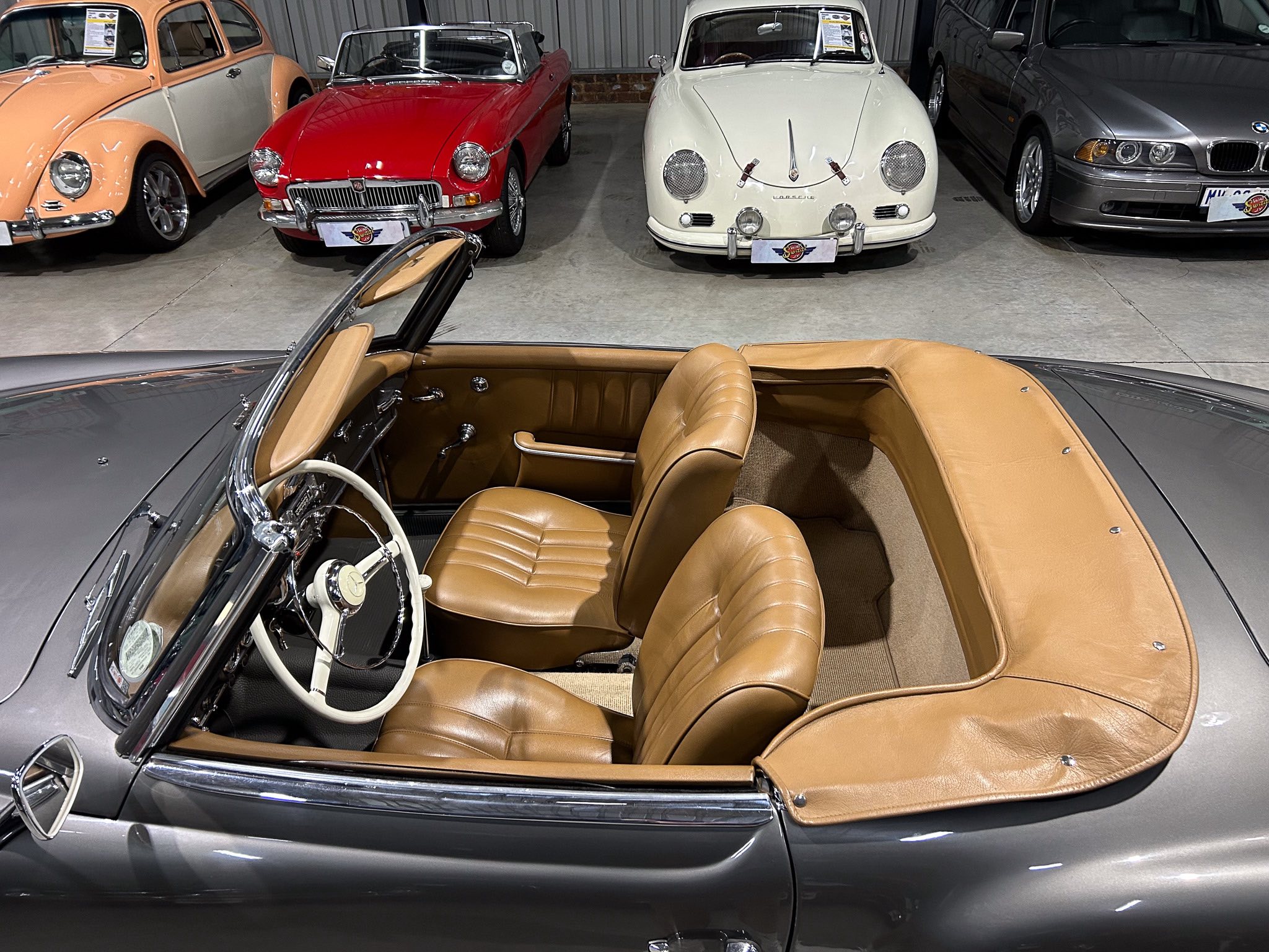 Interior of a vintage car showroom featuring a gray convertible with tan leather interior in the foreground and classic cars parked behind it, including a red convertible and white Porsche.