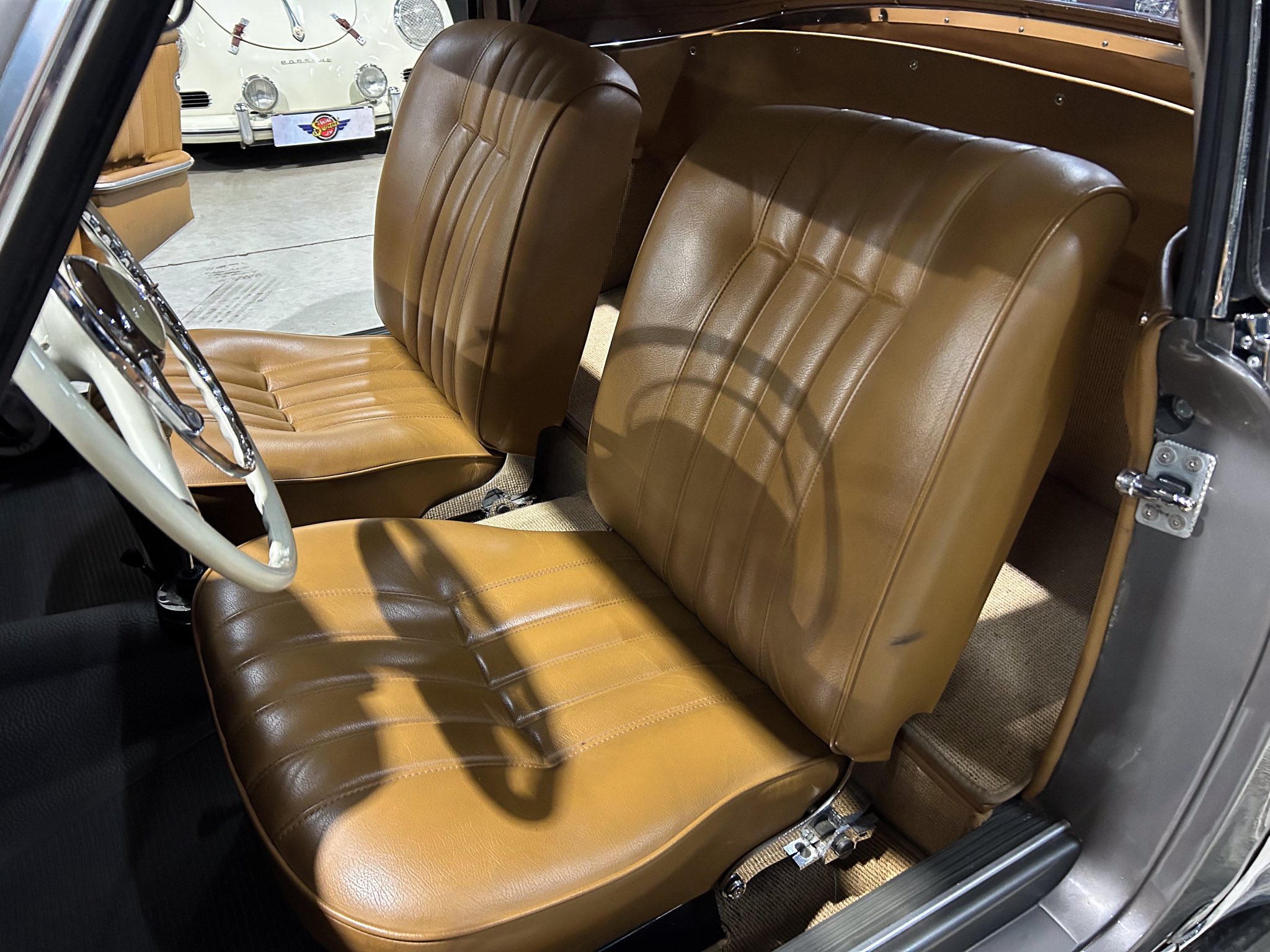 Tan leather front seats of a vintage car interior, with a white classic car visible through the door opening in the background.