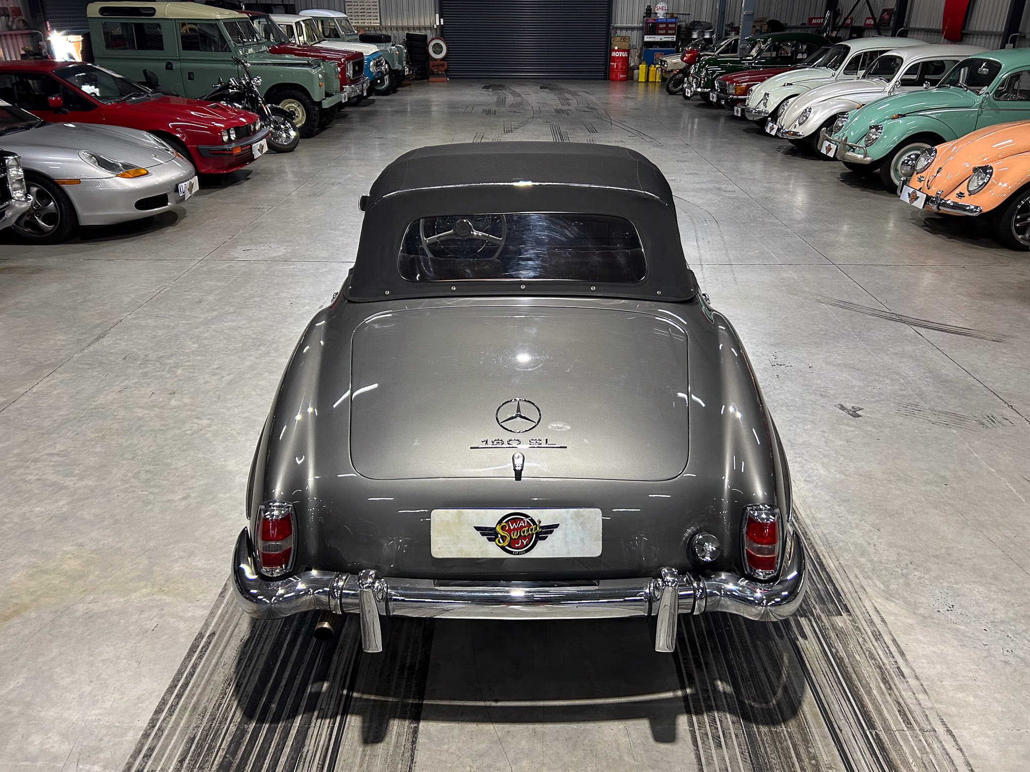 Rear view of a gray Mercedes convertible in a classic car showroom, with vintage cars lined along both walls on either side.