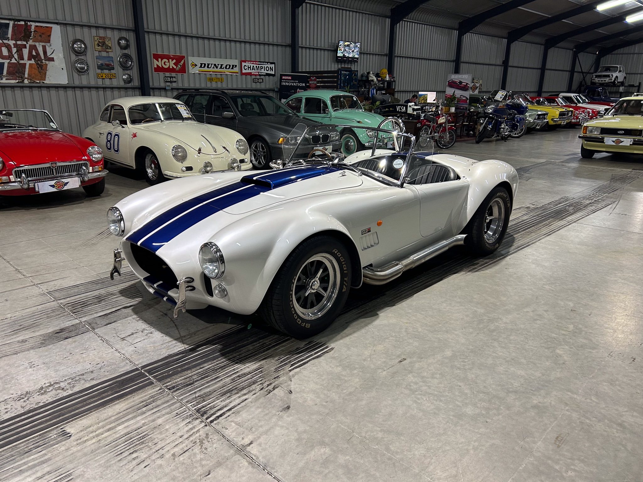 Indoor classic car showroom with multiple vintage cars parked side by side, including a white car with blue racing stripe in the foreground and a gray SUV and pastel green car in the middle, signs on the wall, and a high-ceiling metal building.