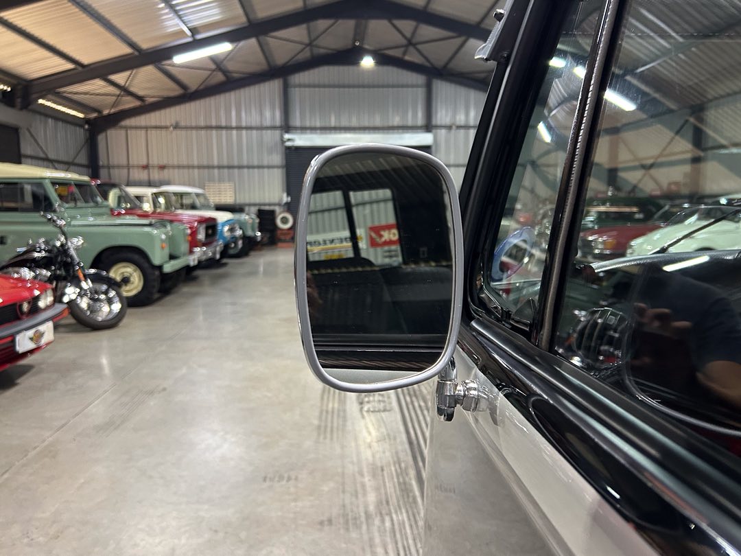 Close-up of a vintage car's side mirror mounted on the driver's side, reflecting the garage interior with classic vehicles lined up inside.