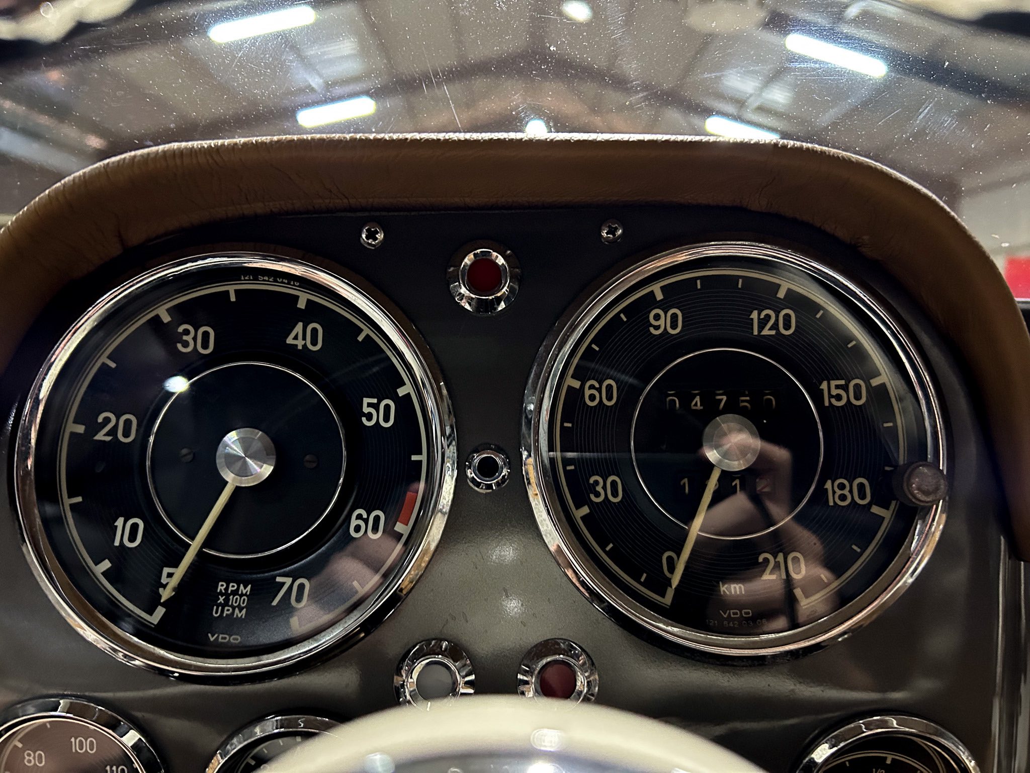 Close-up of a vintage car dashboard with two circular gauges (tachometer on the left, speedometer on the right) surrounded by chrome rings and brown leather trim, and a small red indicator light between them.