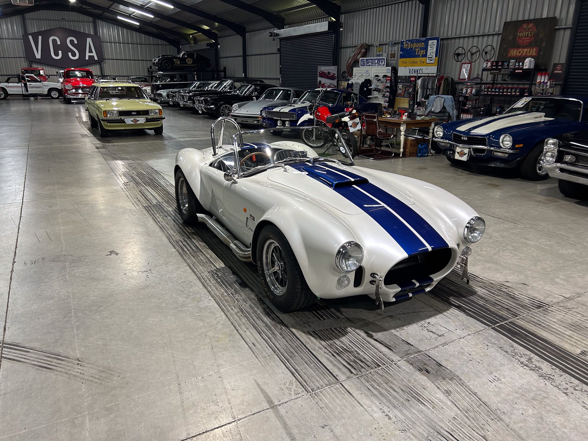White convertible sports car with blue racing stripes in a large indoor classic car showroom. It’s surrounded by vintage cars and automotive memorabilia.