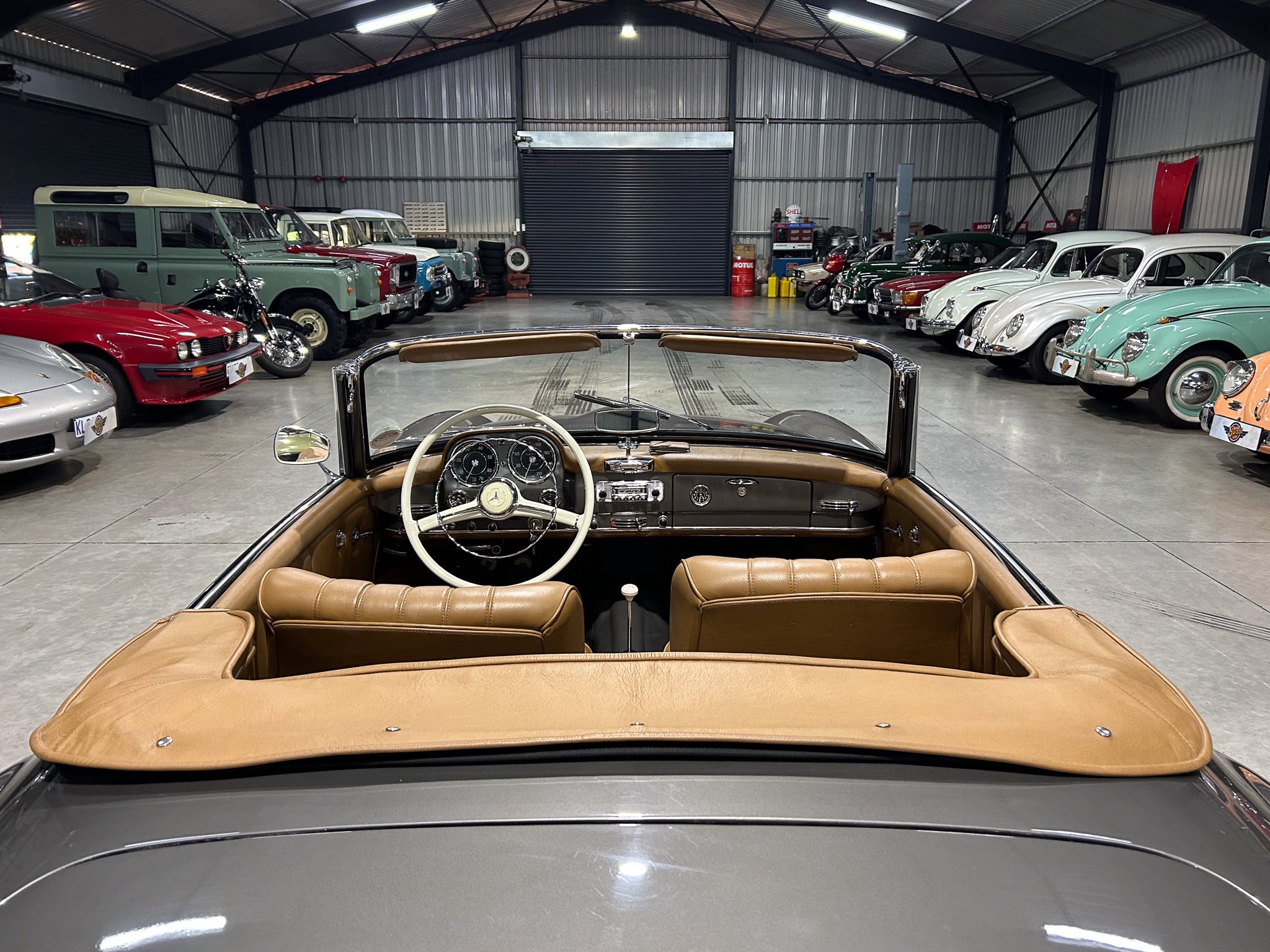 View from the driver's seat of a tan convertible inside a large showroom filled with vintage cars along both sides.
