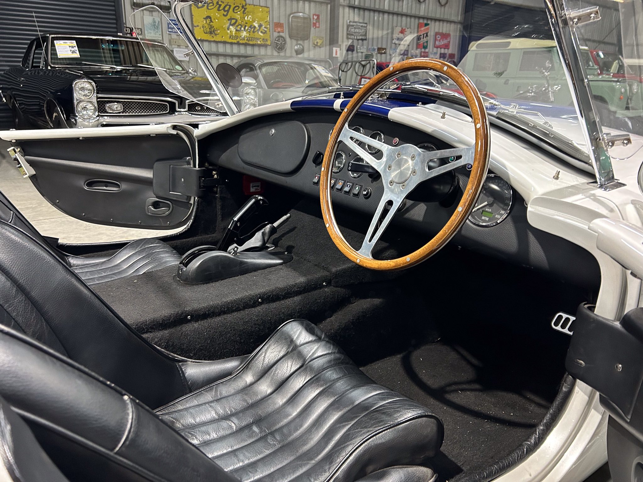 Classic car interior with a wooden steering wheel, black leather seats, and a vintage dashboard inside a showroom garage.
